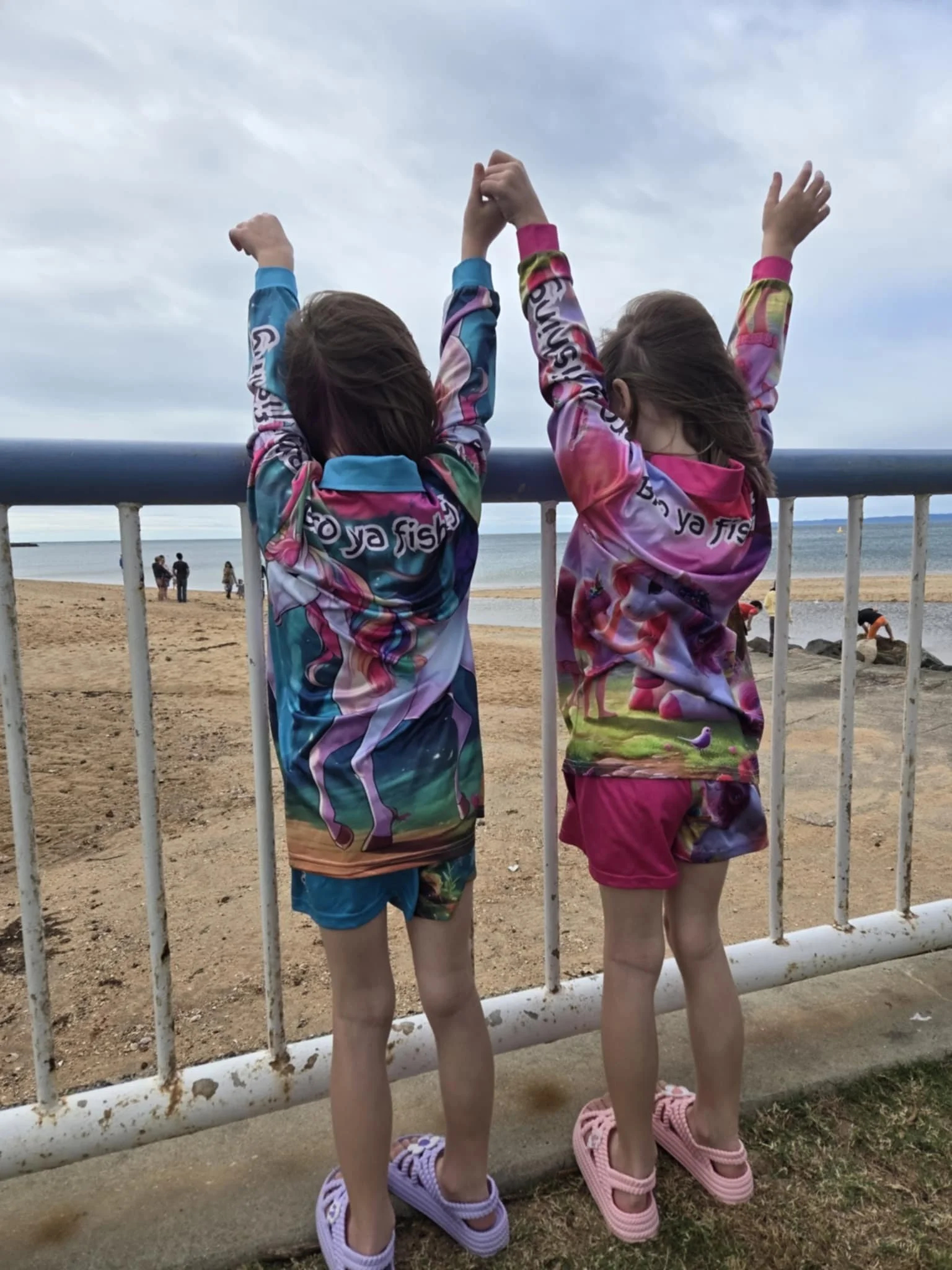 Two children in colorful hoodies and shorts standing by a beach fence, with their backs to the camera, arms raised, overlooking the beach and ocean on a cloudy day.