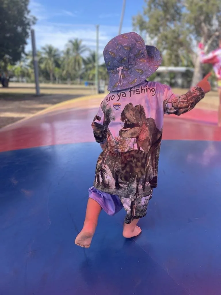 Child wearing a colorful hat and a dinosaur-themed shirt, walking barefoot on a multicolored surface outdoors.