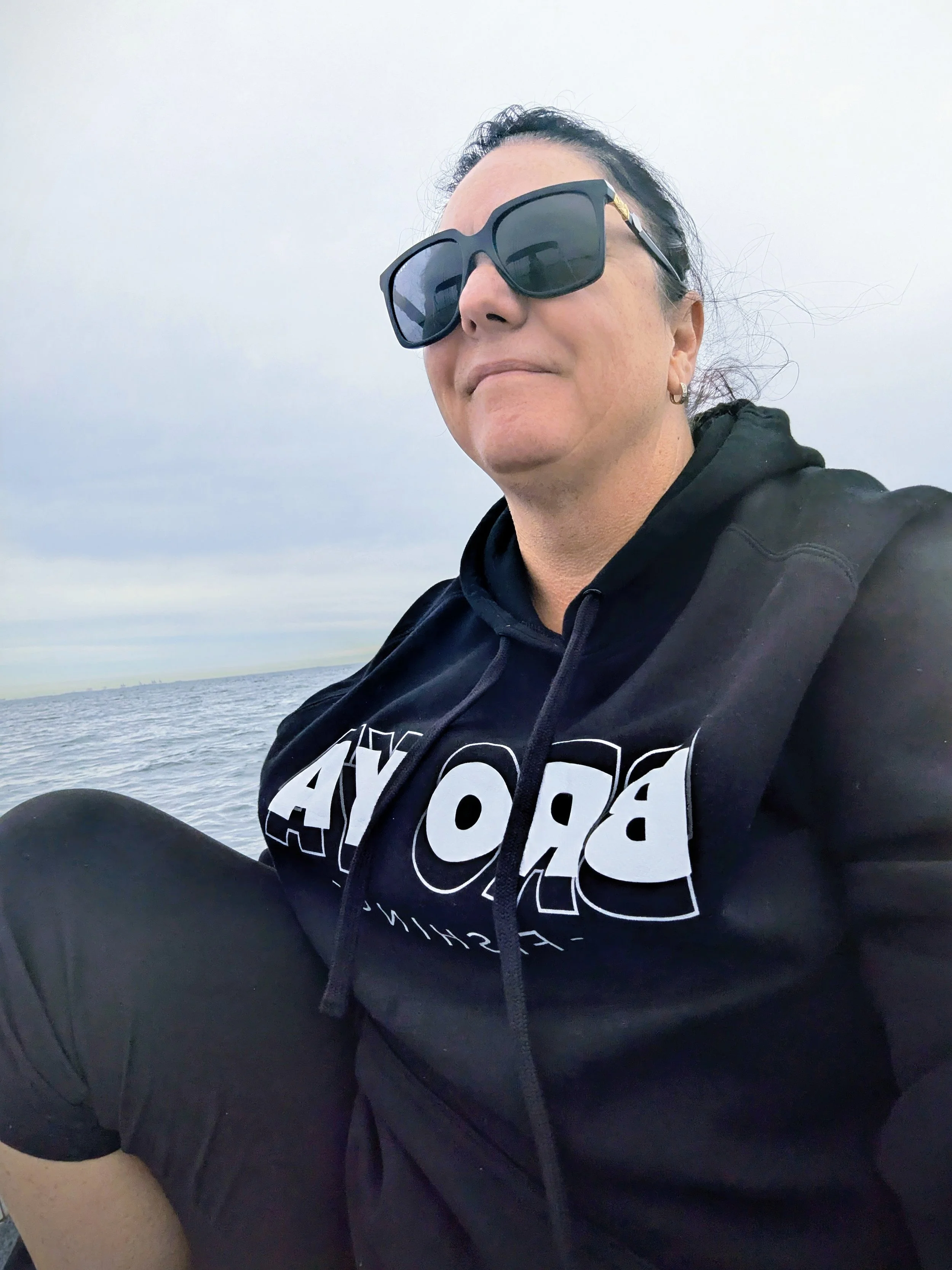 Woman sitting outdoors by water, wearing large sunglasses and a black hoodie with white text, cloudy sky in the background.