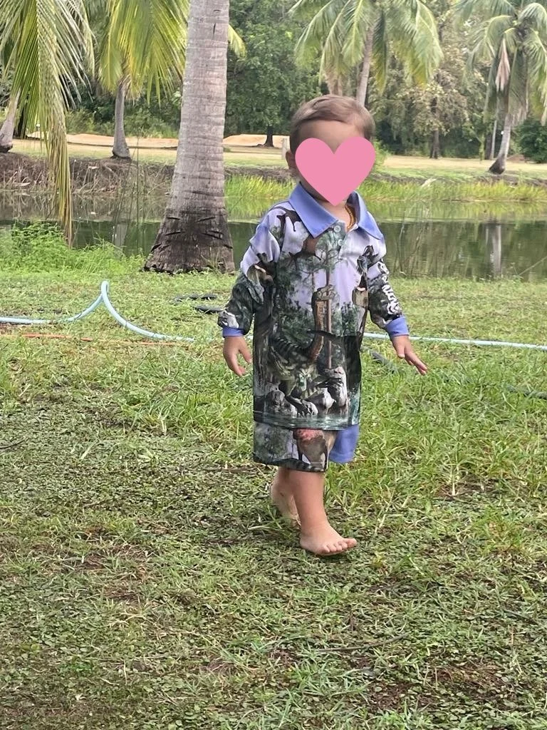 A young child walking barefoot outdoors near a pond, wearing a long-sleeve shirt and shorts with a jungle and animal print, carrying a backpack, with palm trees in the background.