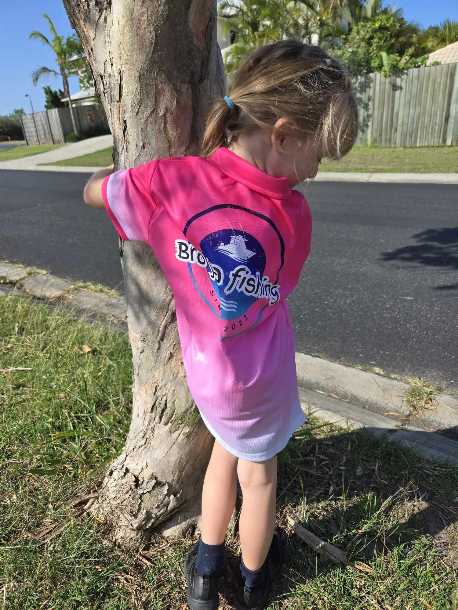 Young girl in a pink tie-dye shirt with a logo that says brook fishing, standing with her back against a tree on a sidewalk in a neighborhood.