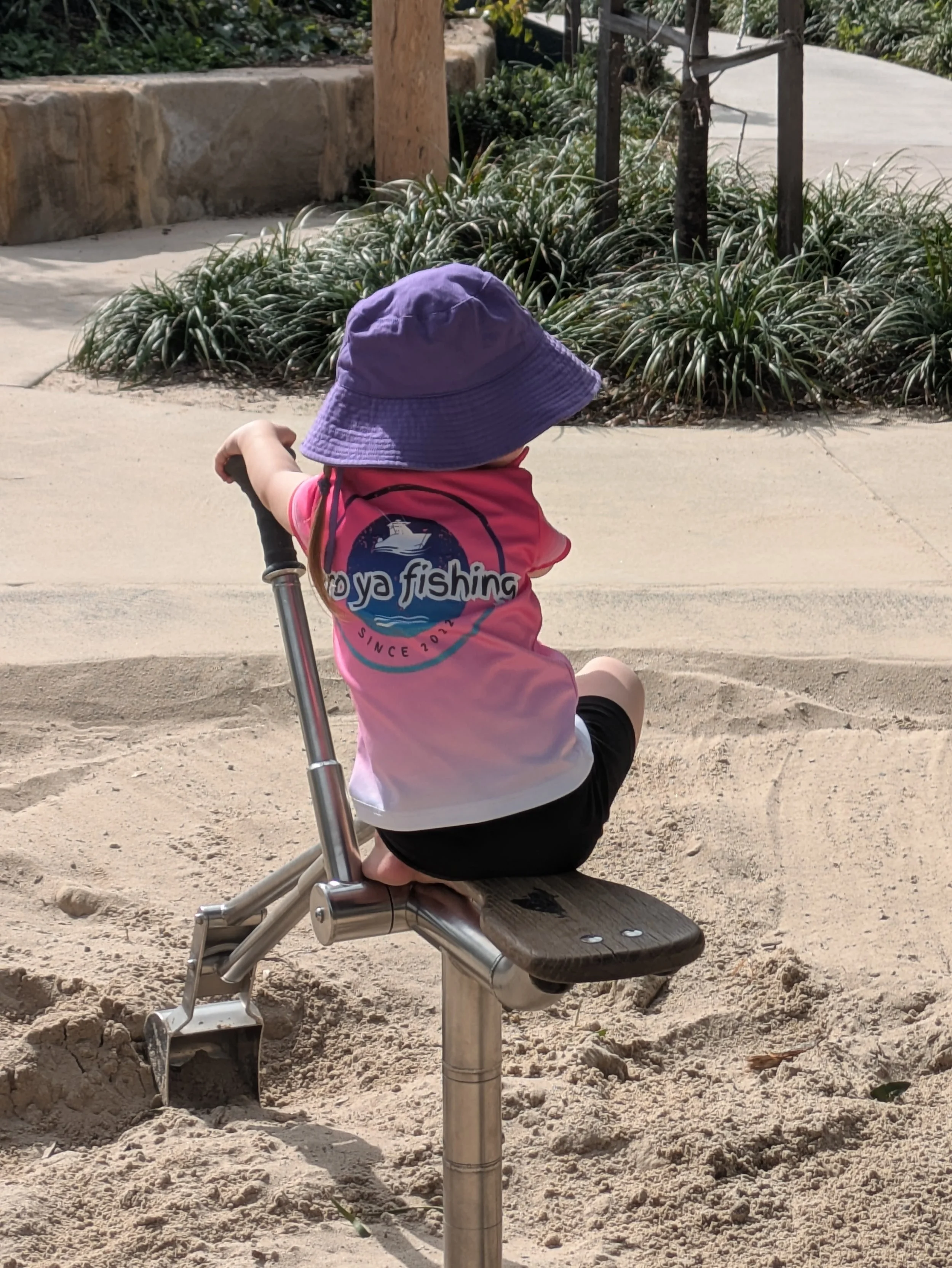 A young child sitting on a small park bench made of wood and metal, wearing a purple sun hat and a pink and white shirt with a boat and fishing graphic, surrounded by sand and greenery.