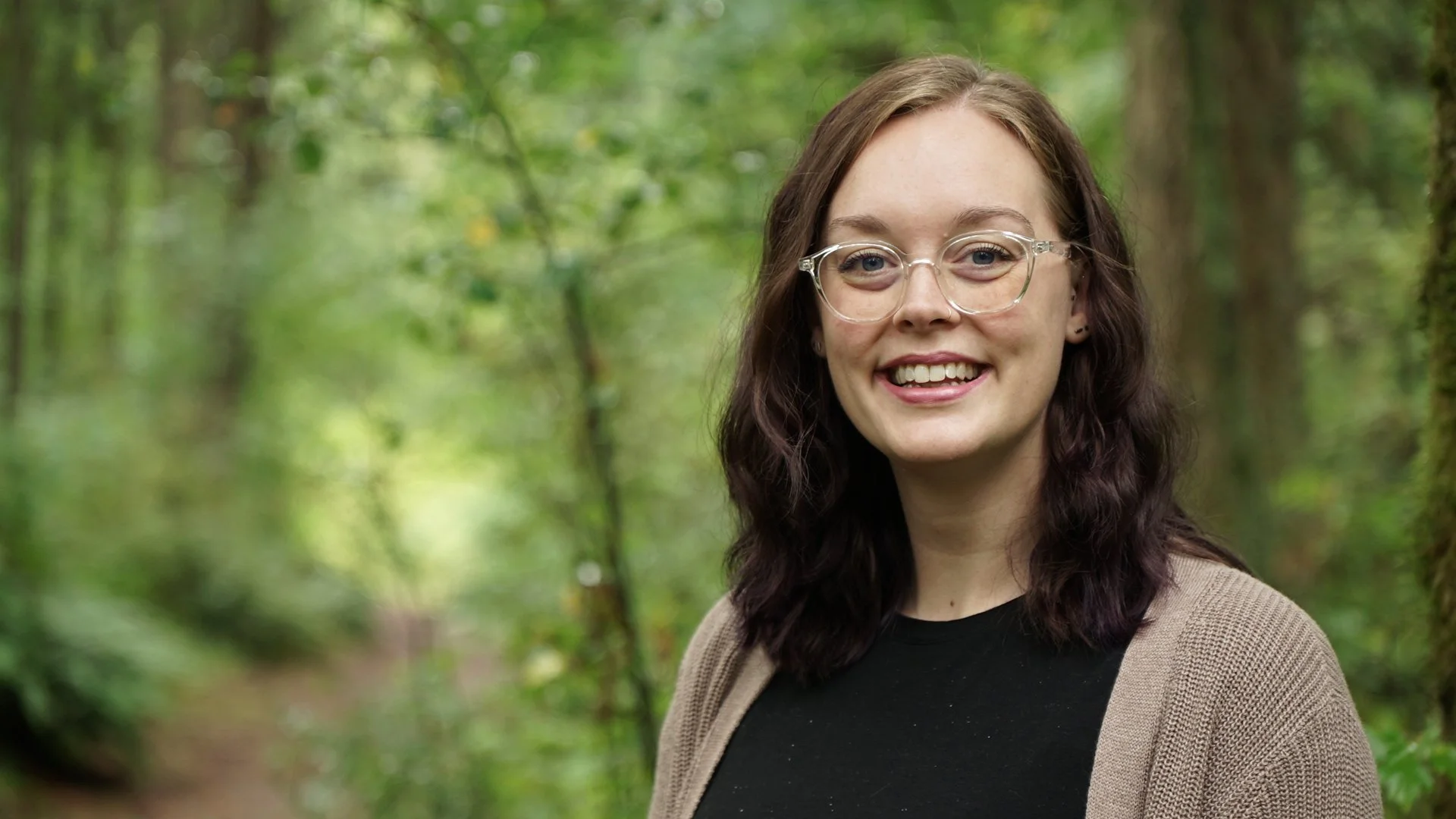 A woman with glasses and long wavy brown hair smiling outdoors in a forest setting.