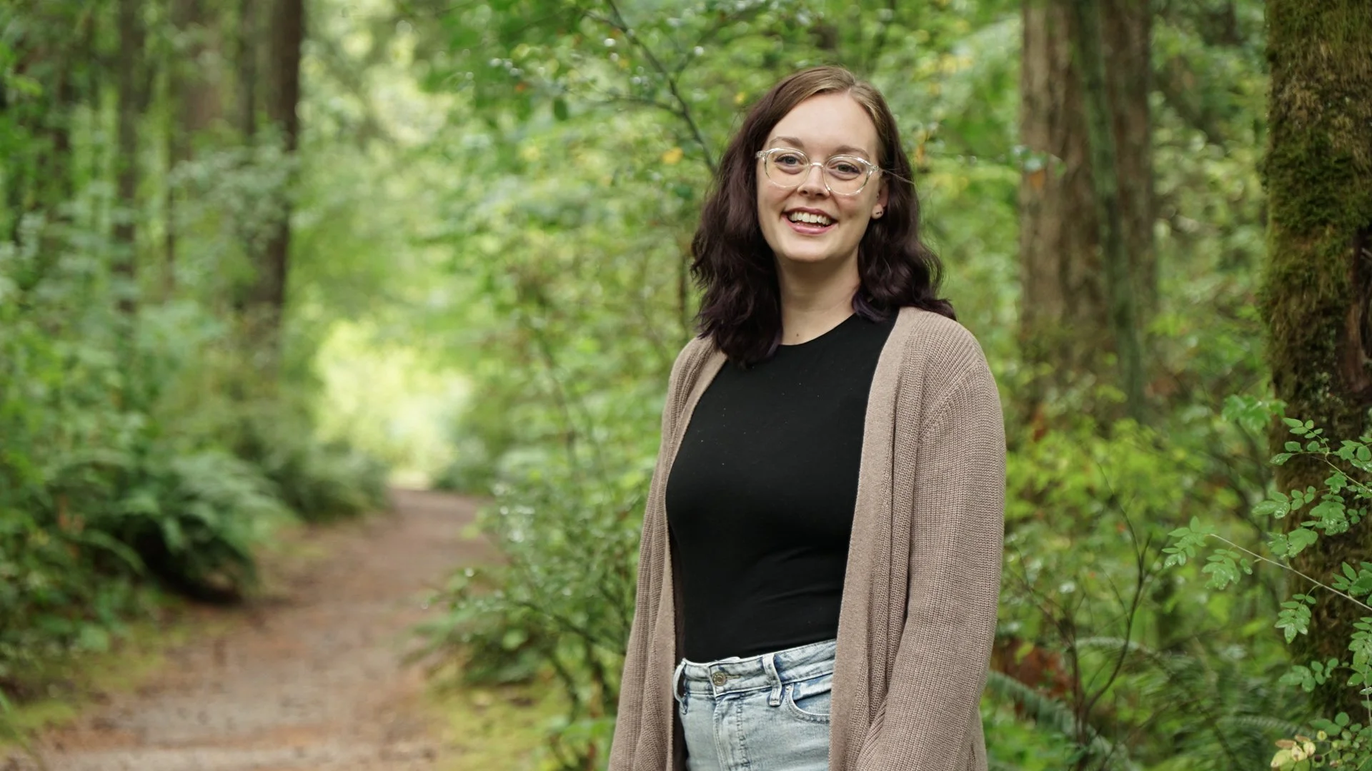 A woman with dark wavy hair, glasses, and a septum piercing smiling in a lush green forest, standing on a dirt path surrounded by trees and foliage.