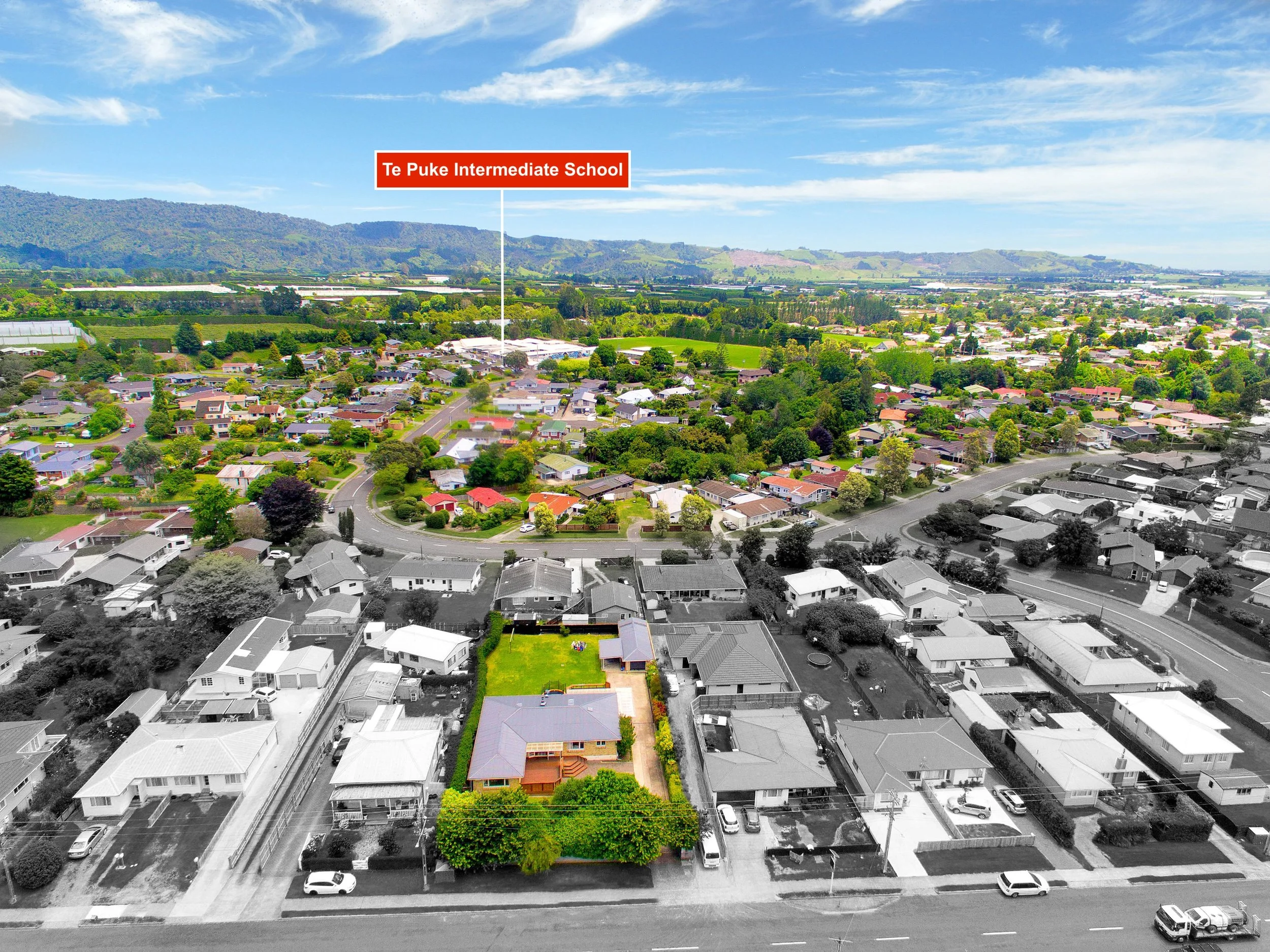Aerial view of a neighborhood with a school labeled 'Te Puke Intermediate School' in the distance. The foreground shows a residential property with a house, trees, and a fenced yard, partly in black and white, with the rest of the neighborhood in col
