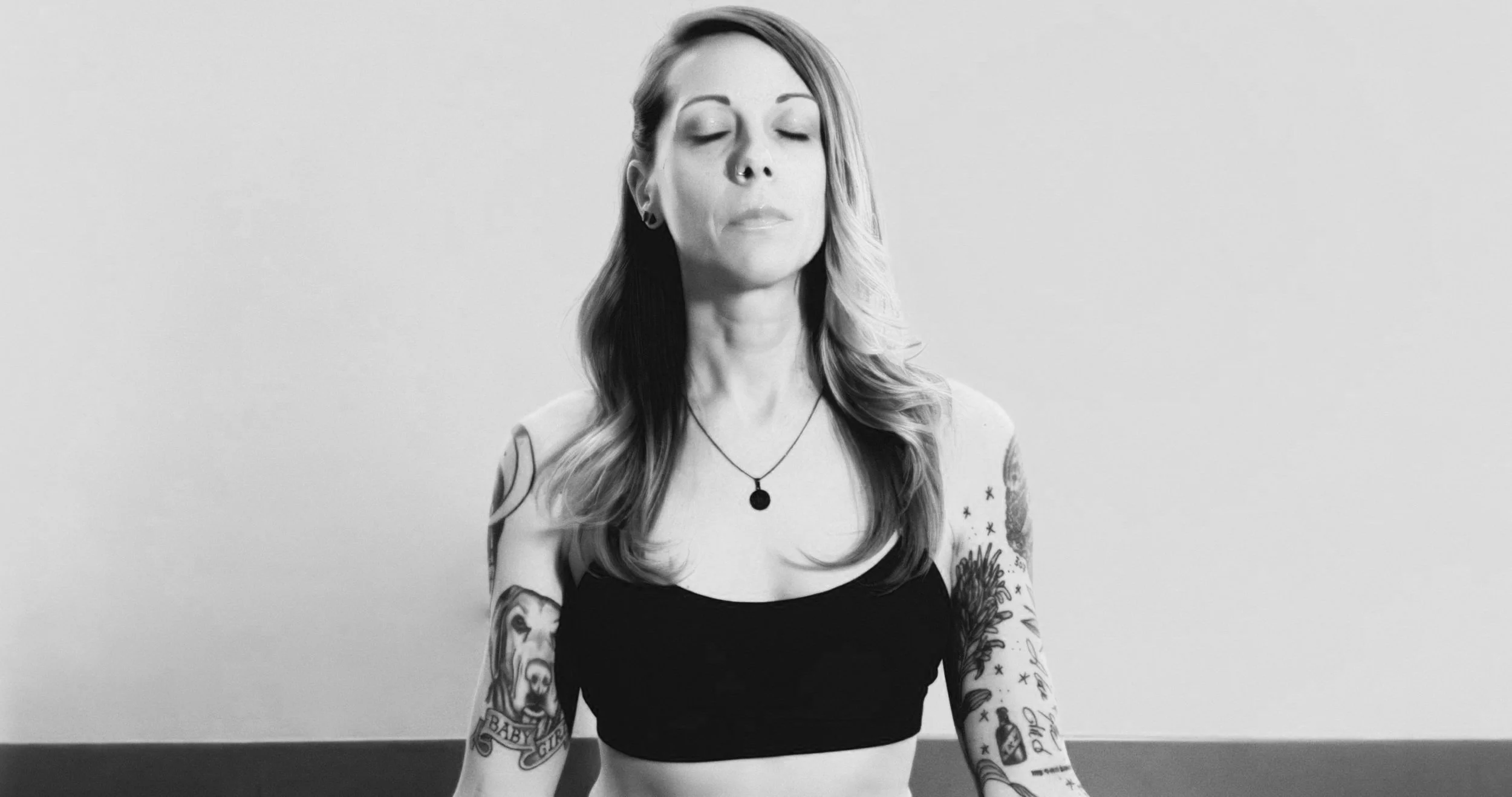 Black and white photo of a woman with tattoos and piercings practicing meditation with her eyes closed, sitting against a plain wall.