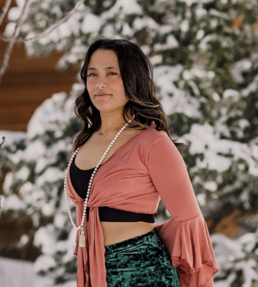 A woman with long dark hair standing outdoors in a snowy landscape with trees in the background, wearing a black crop top, a pink wrap-around top with bell sleeves, a long pearl necklace, and green velvet pants.