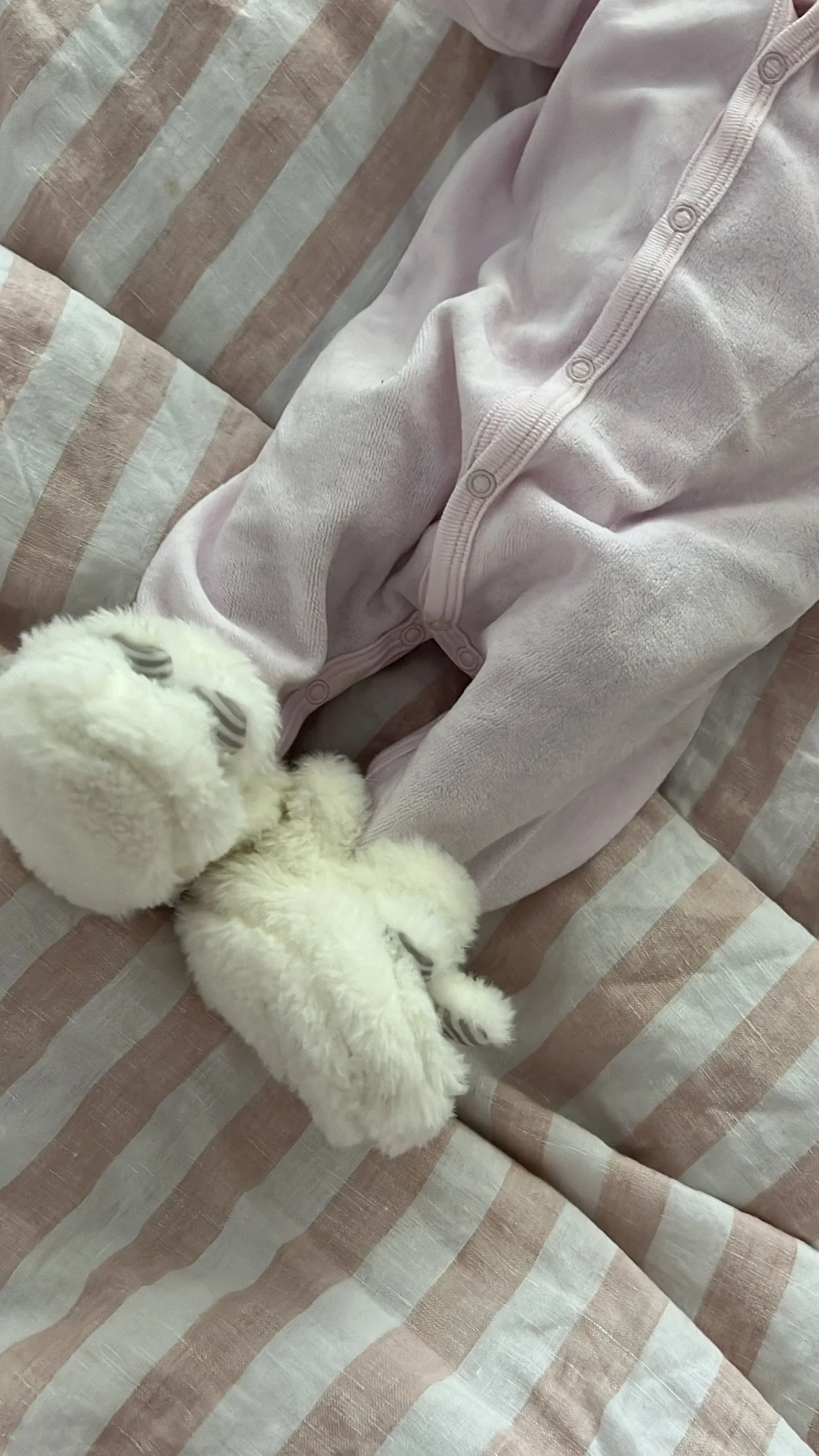 Flat lay of a white baby onesie, pink and green pacifiers, a pink and white baby hat, a gray bow headband, a stuffed teddy bear, on a pink background.