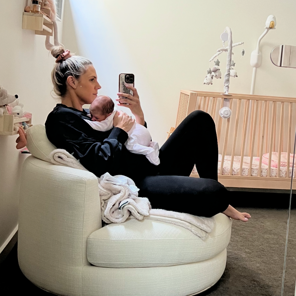 A woman sitting on the carpeted floor in a nursery, holding a newborn baby wrapped in a blanket, with a white crib behind her and books on wall shelves.