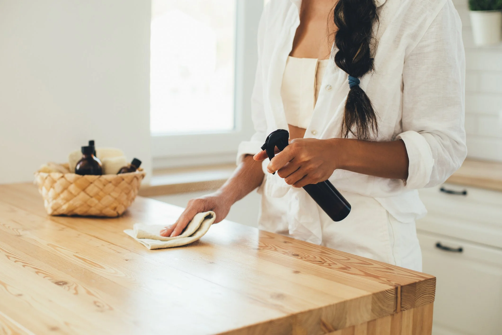 A woman in a white shirt cleaning a wooden kitchen counter with a spray bottle and cloth.