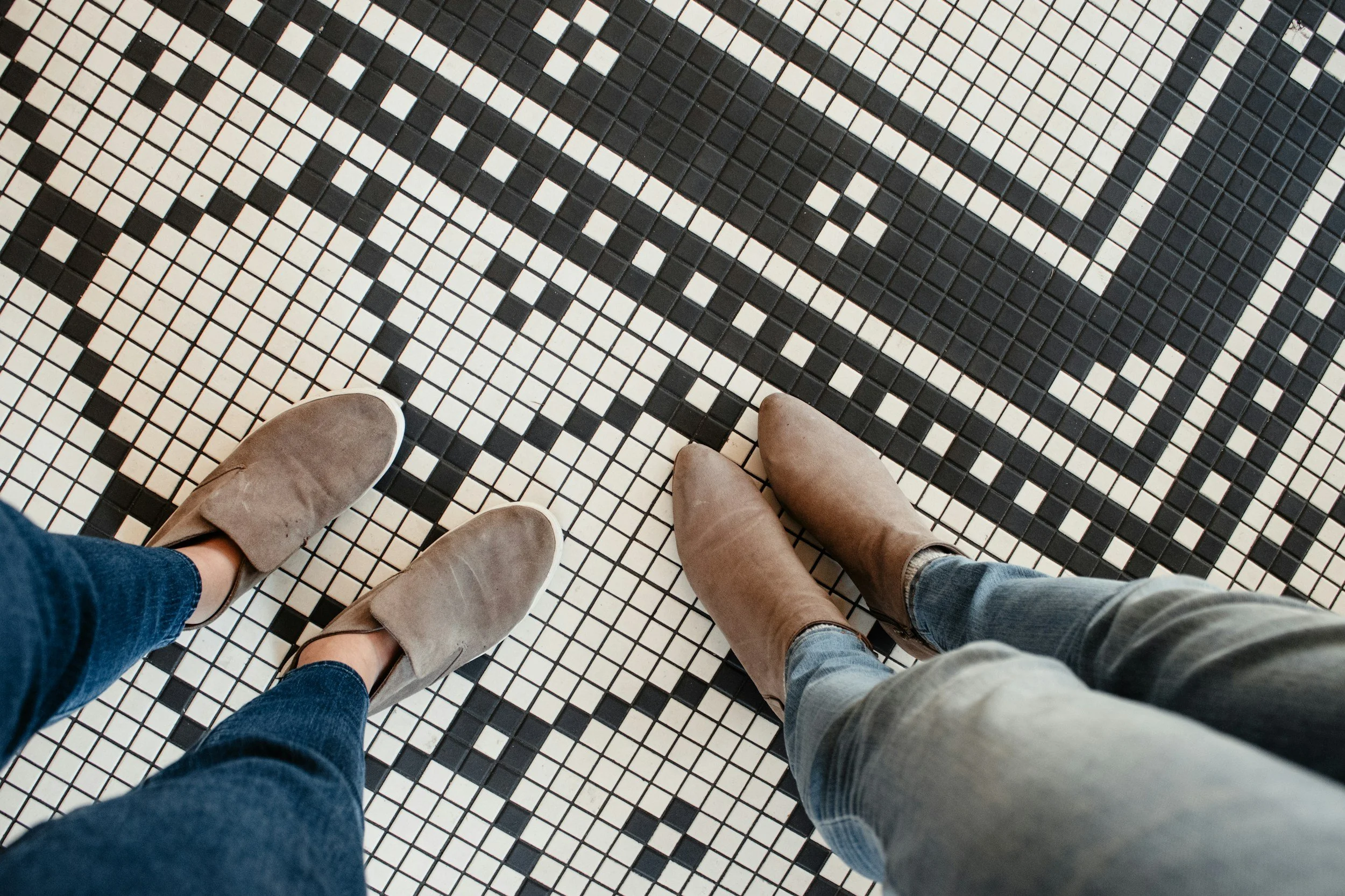 Looking down at two pairs of shoes standing on a black-and-white tiled floor.
