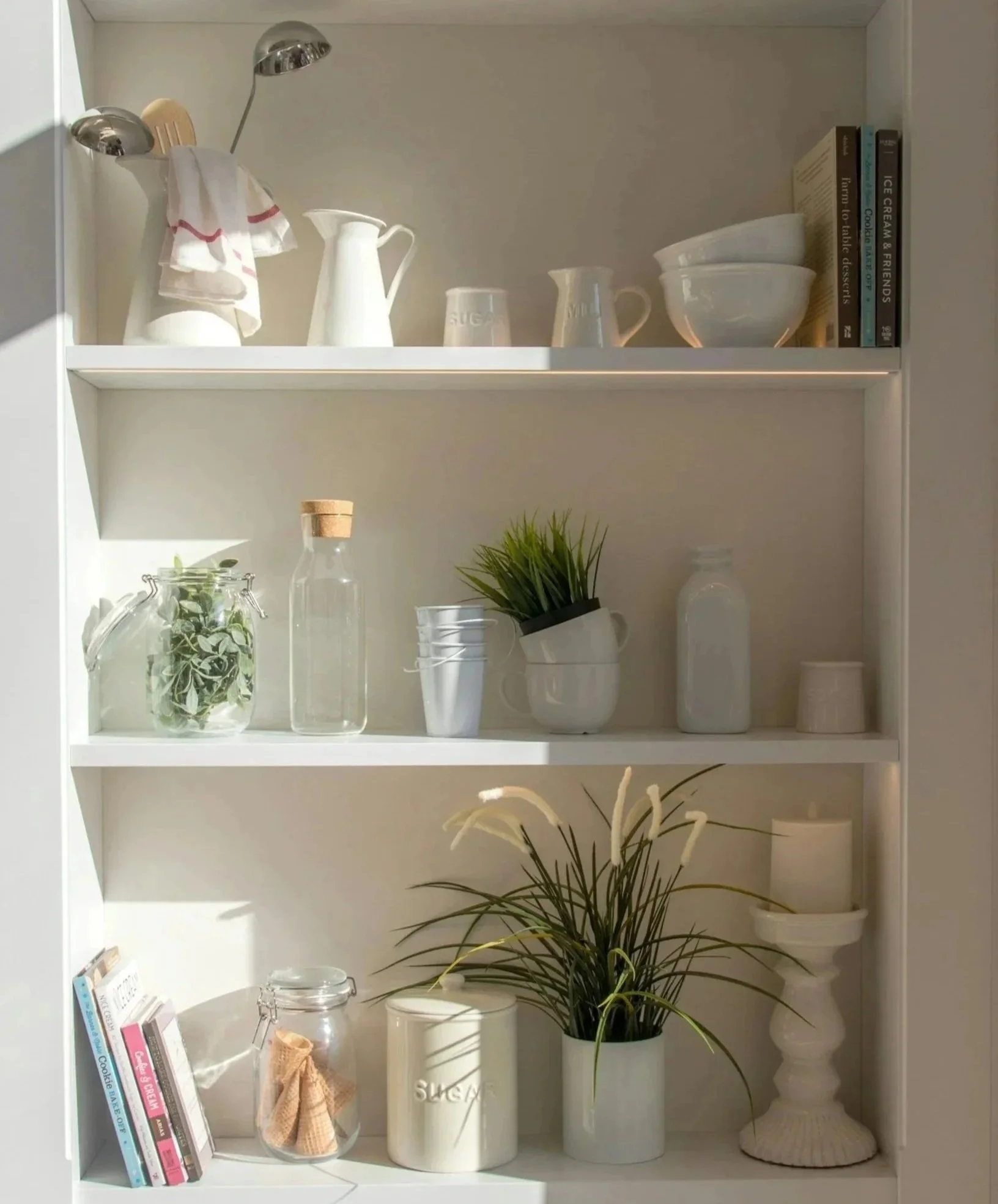 Open white bookshelf with decorative kitchen and plant items, including books, a glass jar with a bow, a white pitcher, a small potted plant, and ceramic baskets. Organization and home staging.