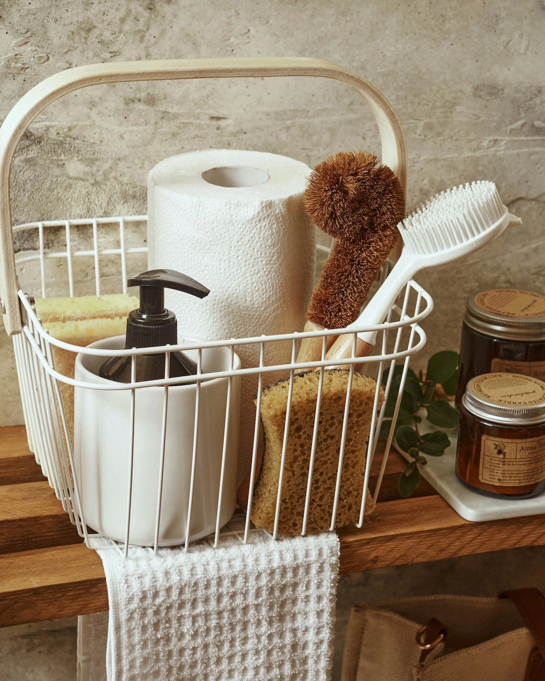 A white wire basket on a wooden surface containing a roll of paper towels, a brown textured scrub brush, a white bristled cleaning brush, a small black spray bottle, and a sponge. In the background, there are jars on a white tray and some greenery.