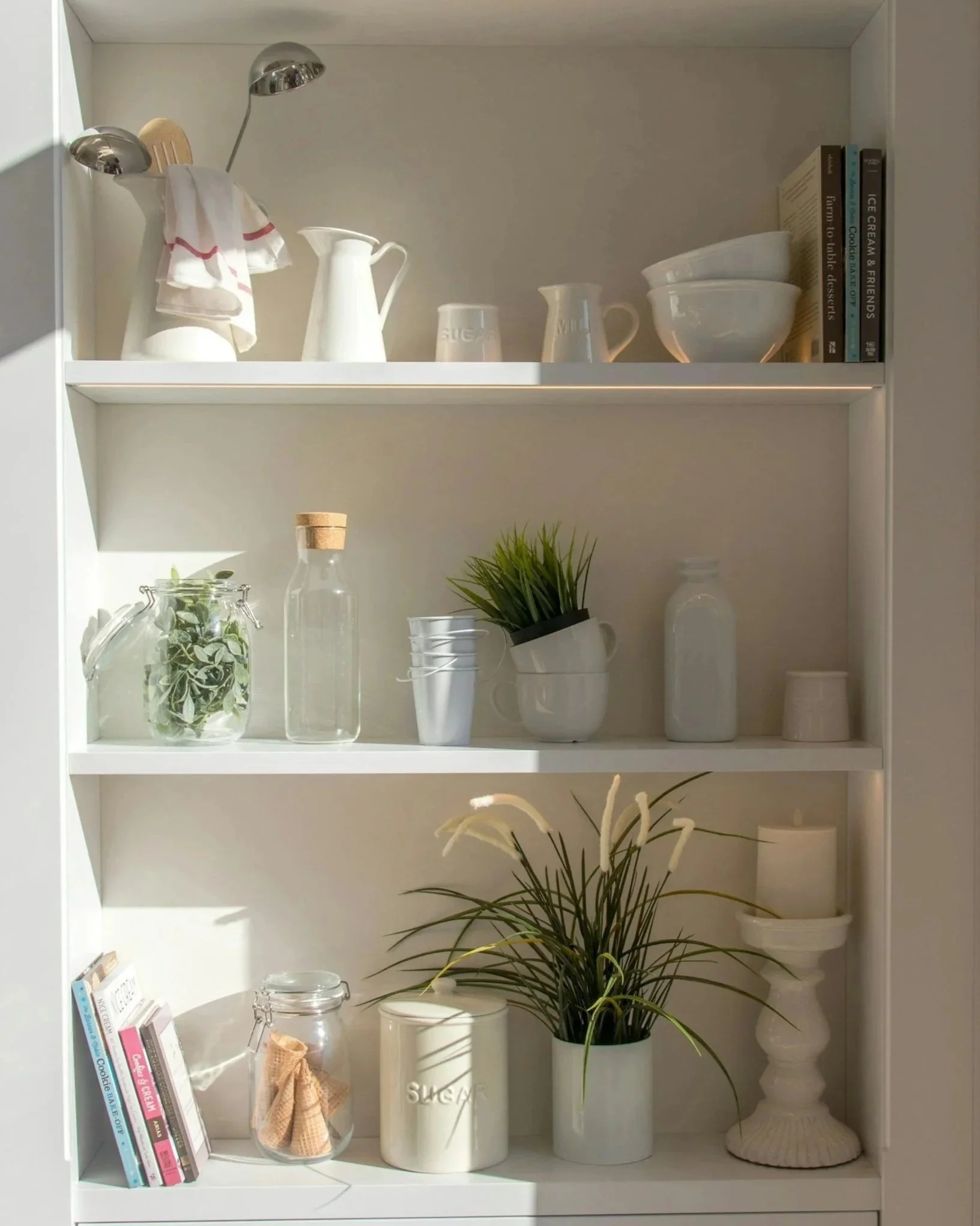 Open white bookshelf with decorative items, cookbooks, and plants. Top shelf has dishes and books, middle has glass jars, a plant, and a small vase, bottom has additional books and a large ceramic vase.
