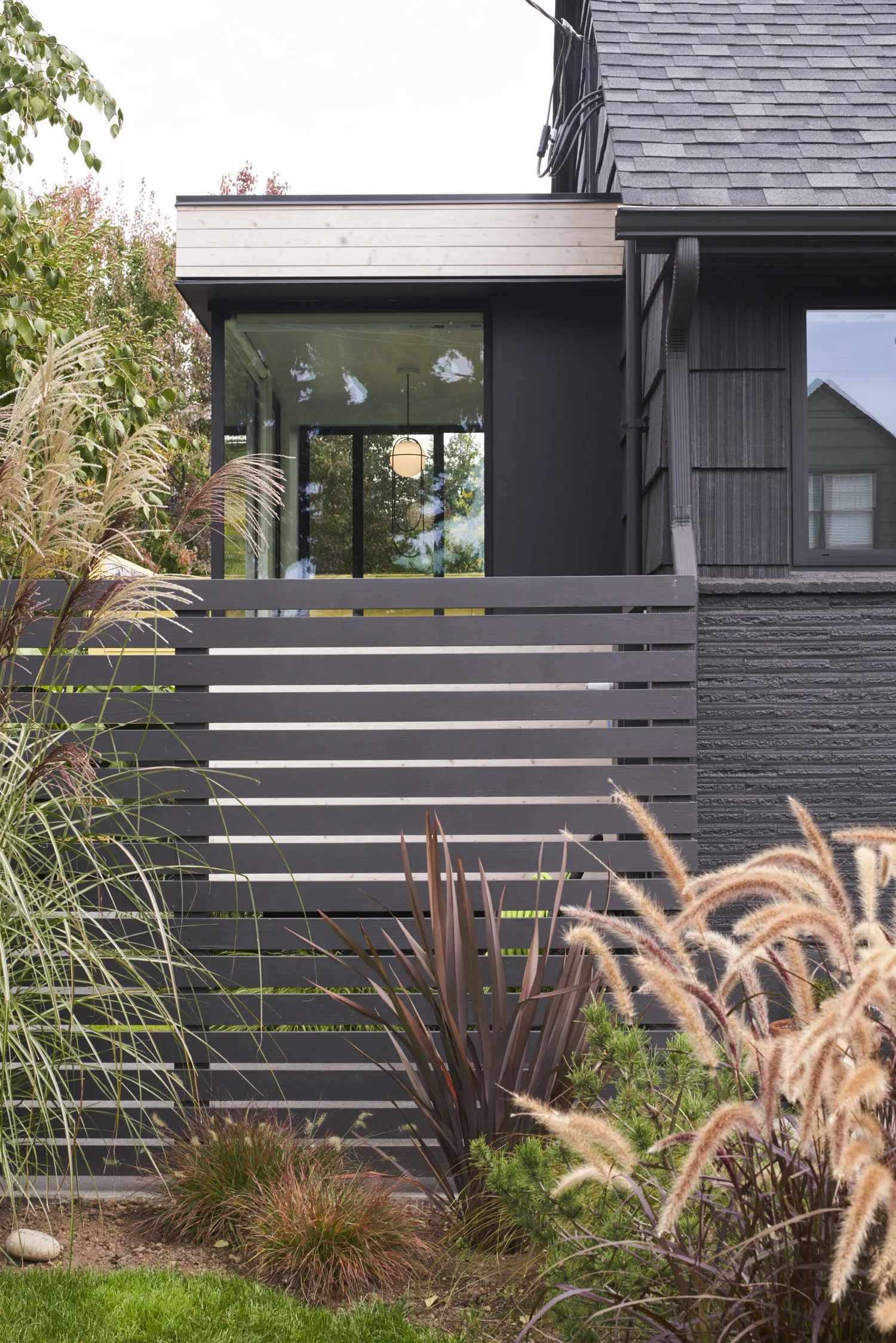 Modern house with black exterior, large glass window, gray horizontal fence, and landscaped garden with ornamental grasses and plants.