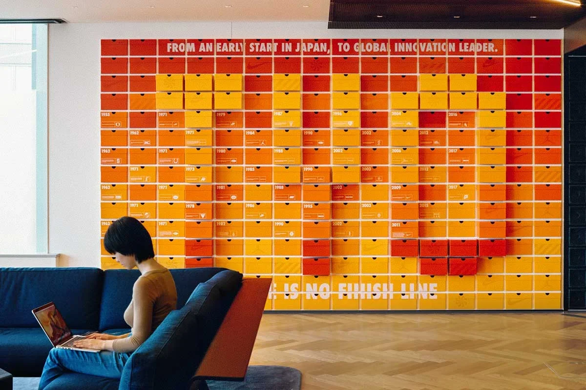 A woman sitting on a dark blue couch using a laptop in front of a colorful wall display that resembles a large calendar or timeline with orange, red, and yellow compartments, each marked with years and symbols.
