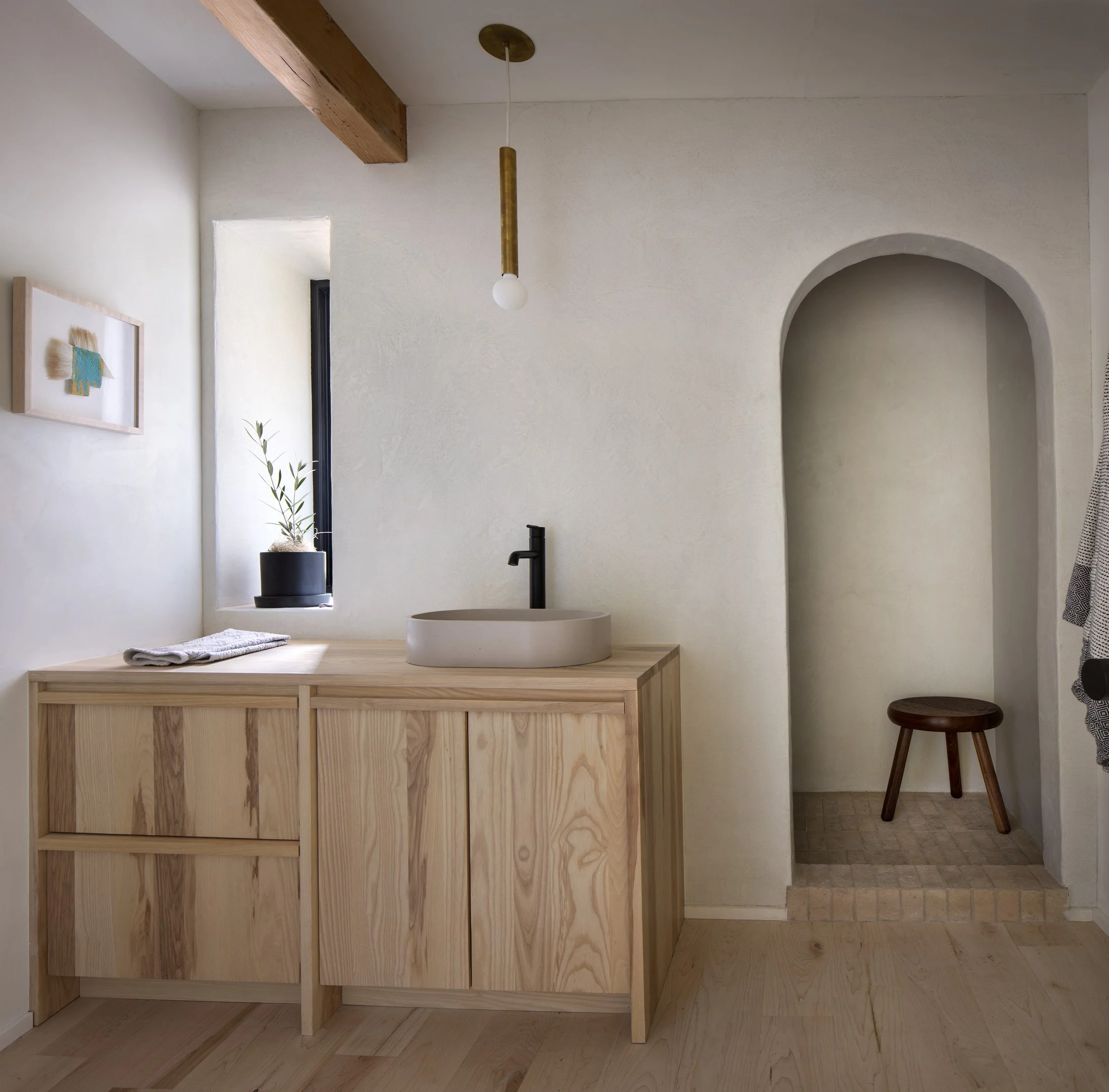 Minimalist bathroom with wooden vanity, white vessel sink, black faucet, small window with a plant, framed artwork, hanging light, and an arched alcove with a small wooden stool.