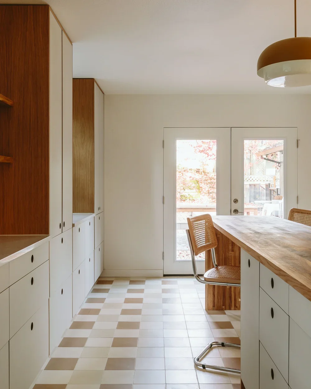 A modern kitchen with white and wood cabinetry, tiled floor, and a large wooden kitchen island with a chair. Double glass doors lead outside to a deck, with trees visible through the glass.