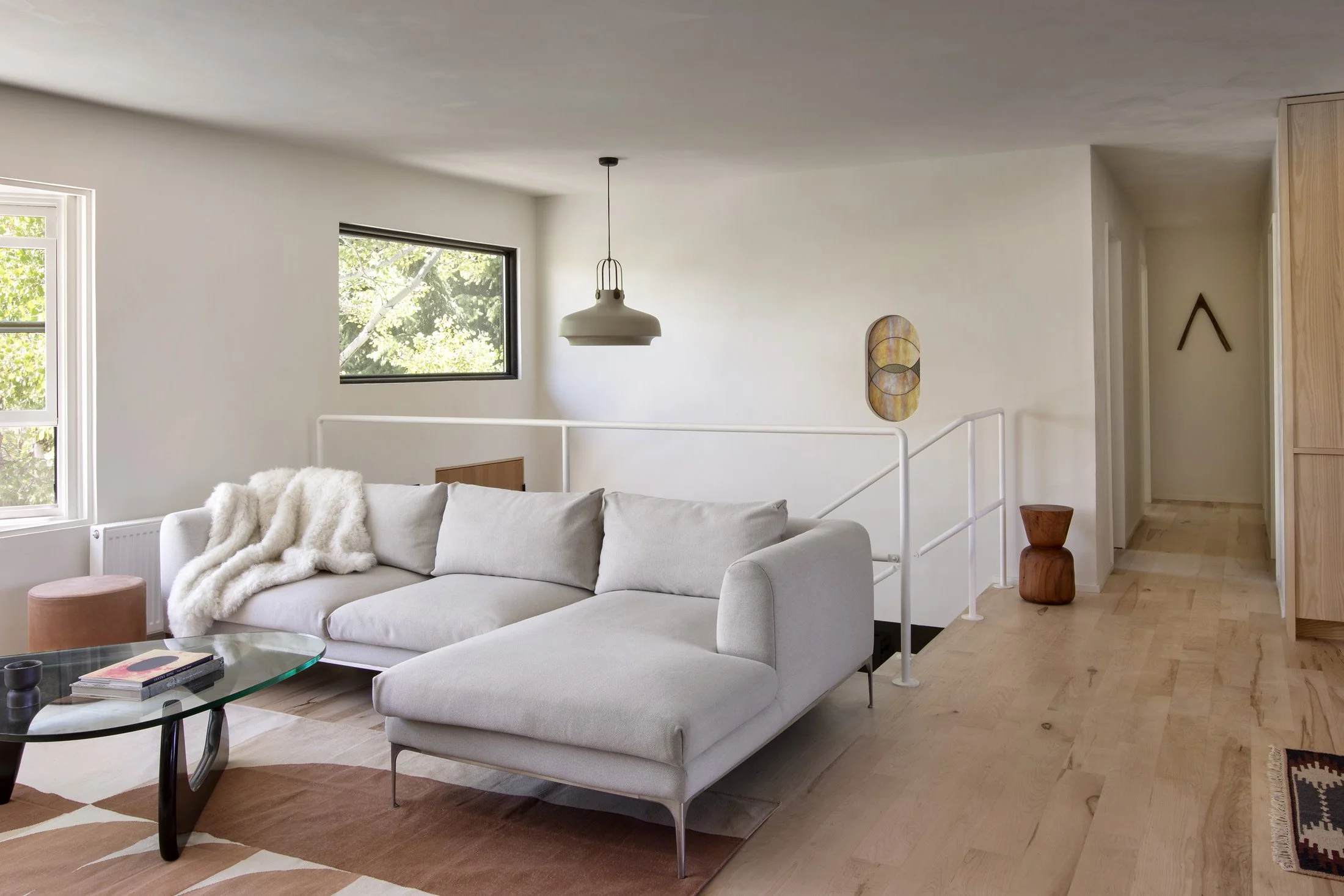 Minimalist living room with white sofa, glass coffee table, wooden side table, wall art, and large windows showing greenery outside.