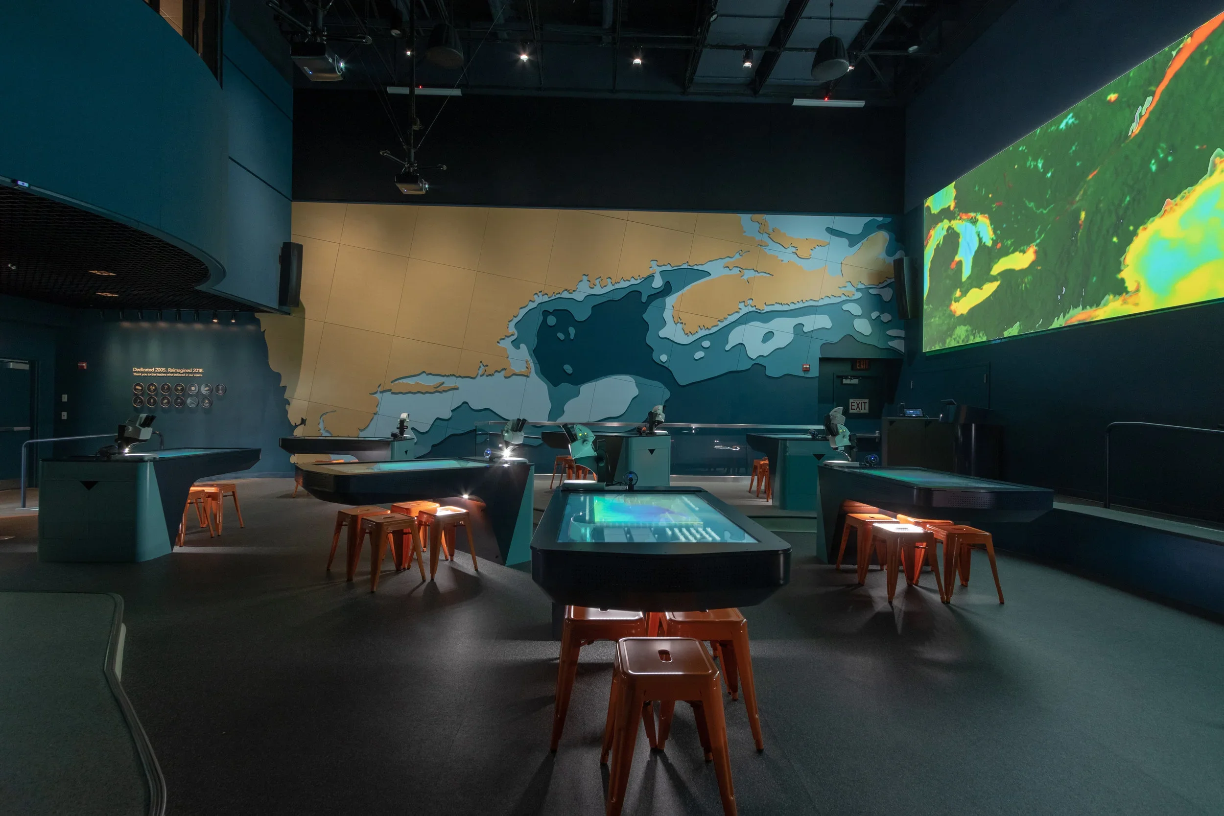 An indoor space with several tables equipped with microscopes, orange stools, and a large wall map of the Great Lakes on the back wall. A large digital screen displaying colorful visuals is on the right side.