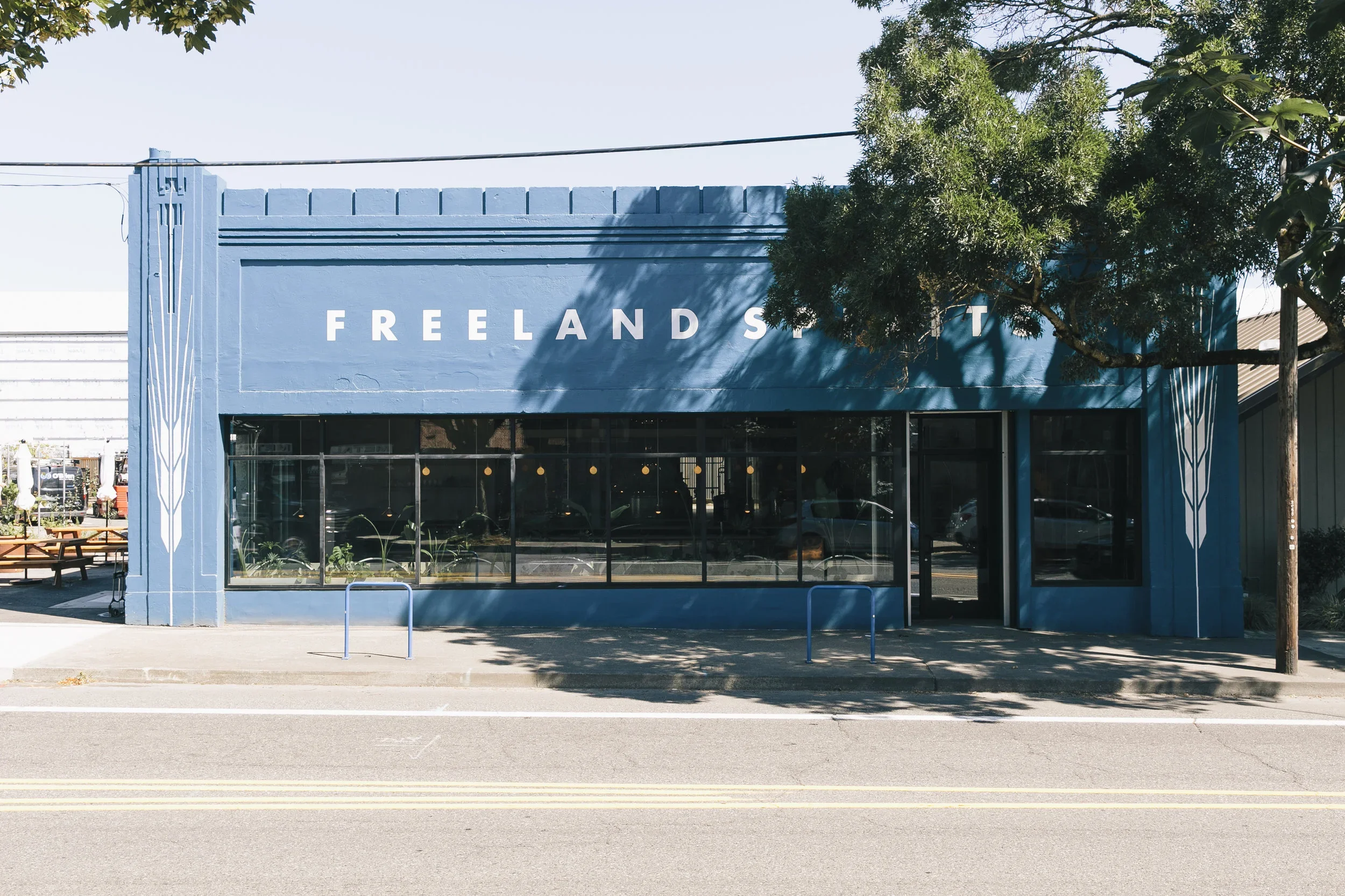 Blue building with the word 'FREELAND STREET' on the front, trees casting shadows, and a sidewalk with bike racks in front.