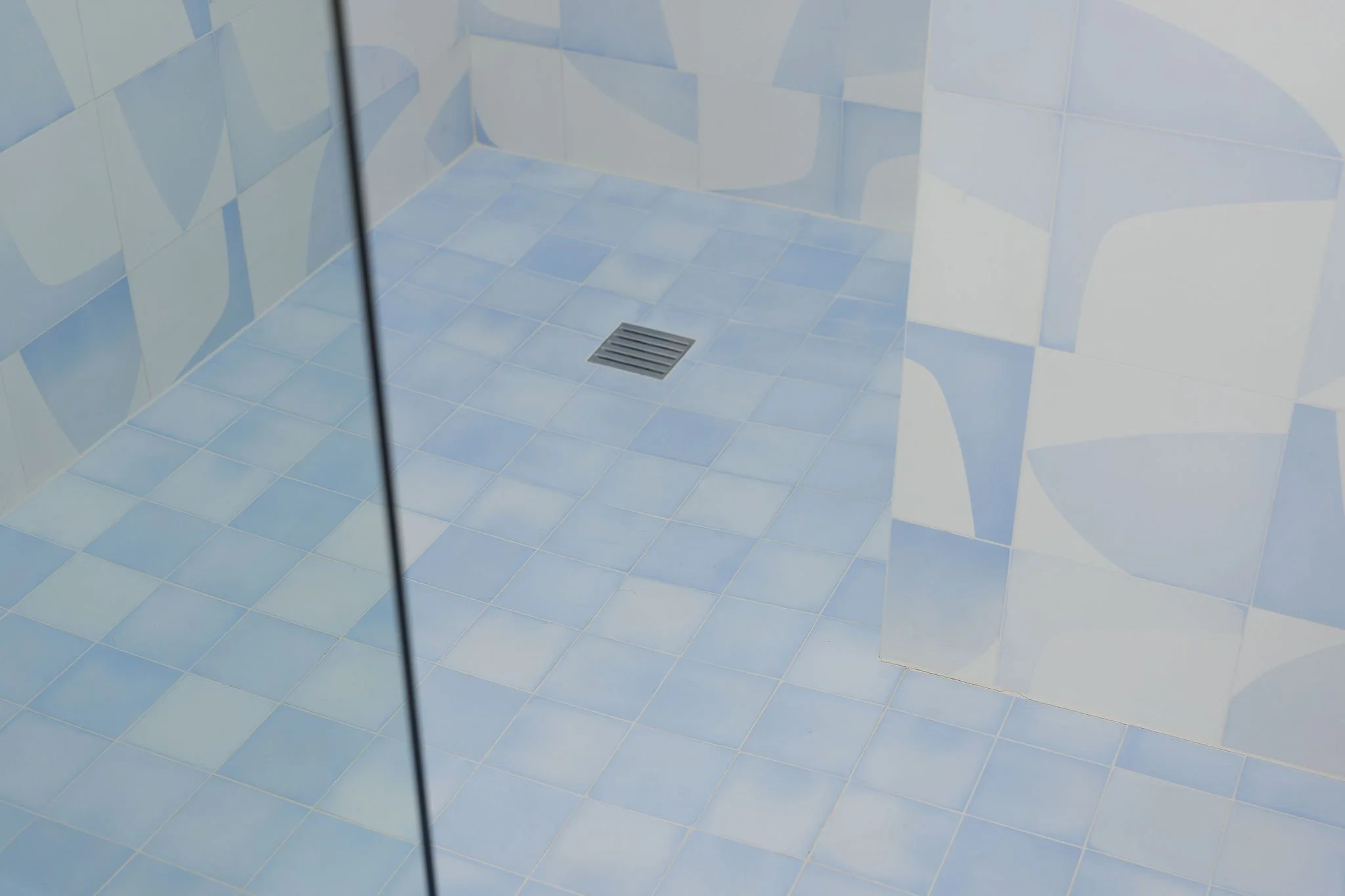 A clean shower area with light blue tiled walls and floor, a square drain in the center, and a mirror reflecting part of the tiled wall.