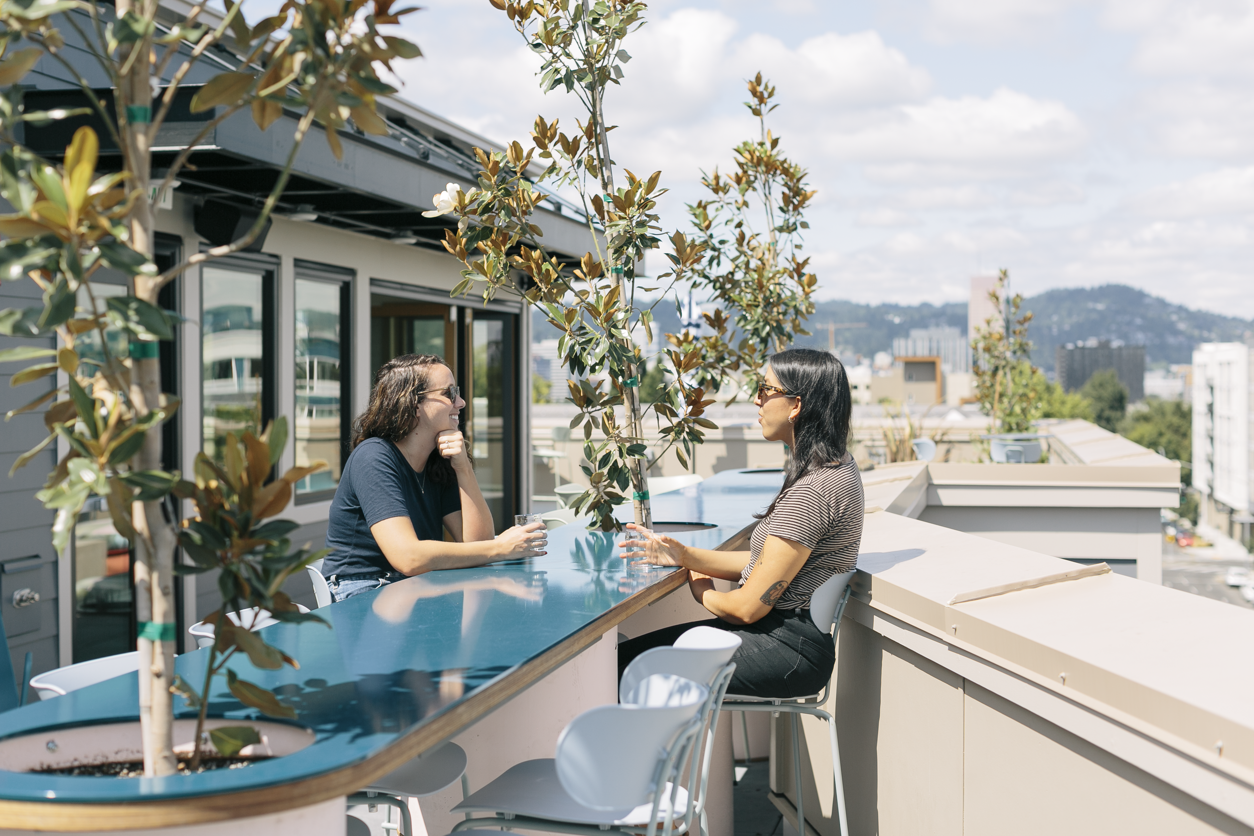 Two women having a conversation at a rooftop bar on a sunny day with a cityscape background, one with glasses and dark hair, the other with head tilt and light brown hair.