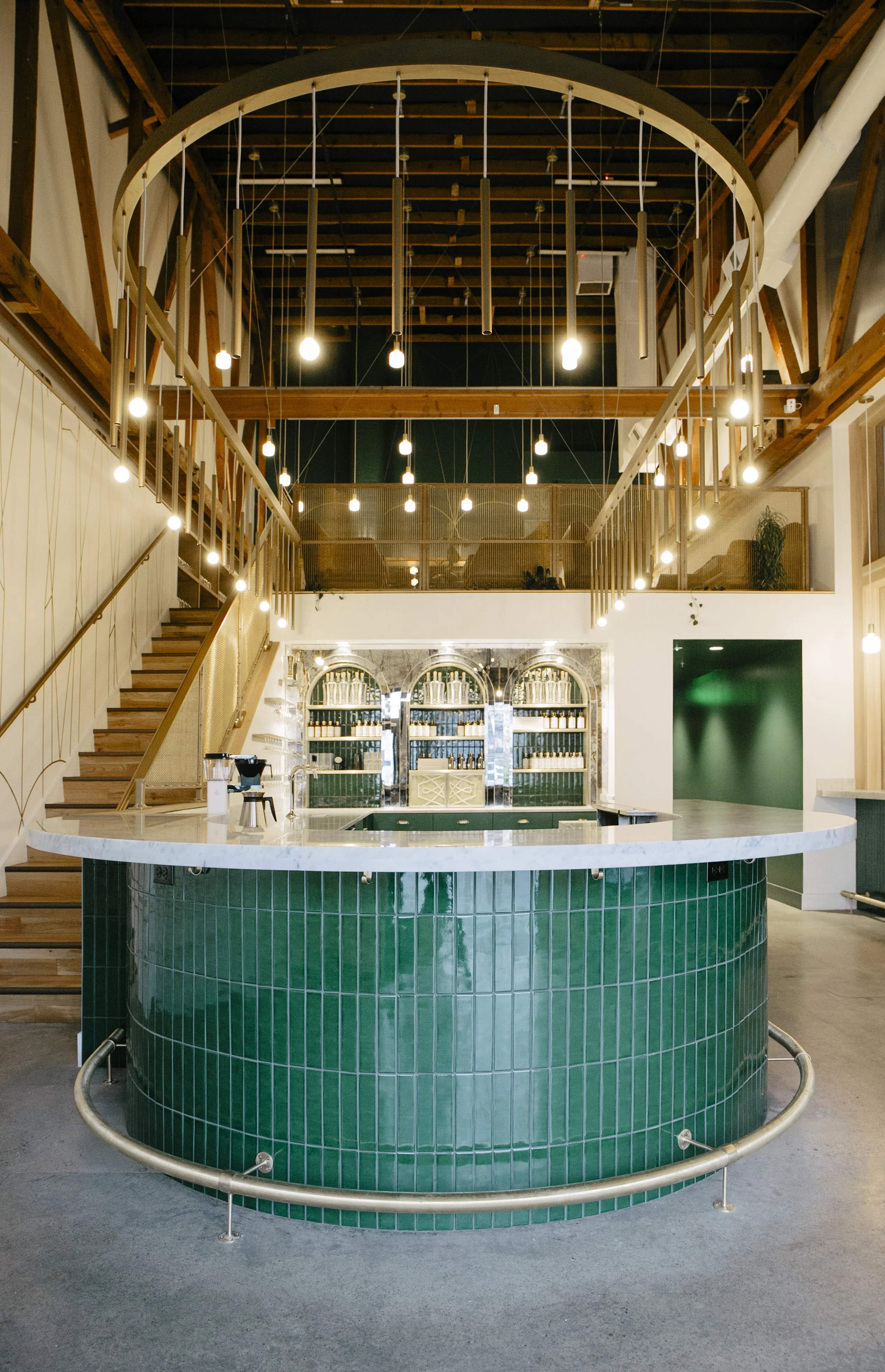 Modern interior of a bar with a green tiled curved counter, marble top, wooden staircase, and hanging Edison-style lights.