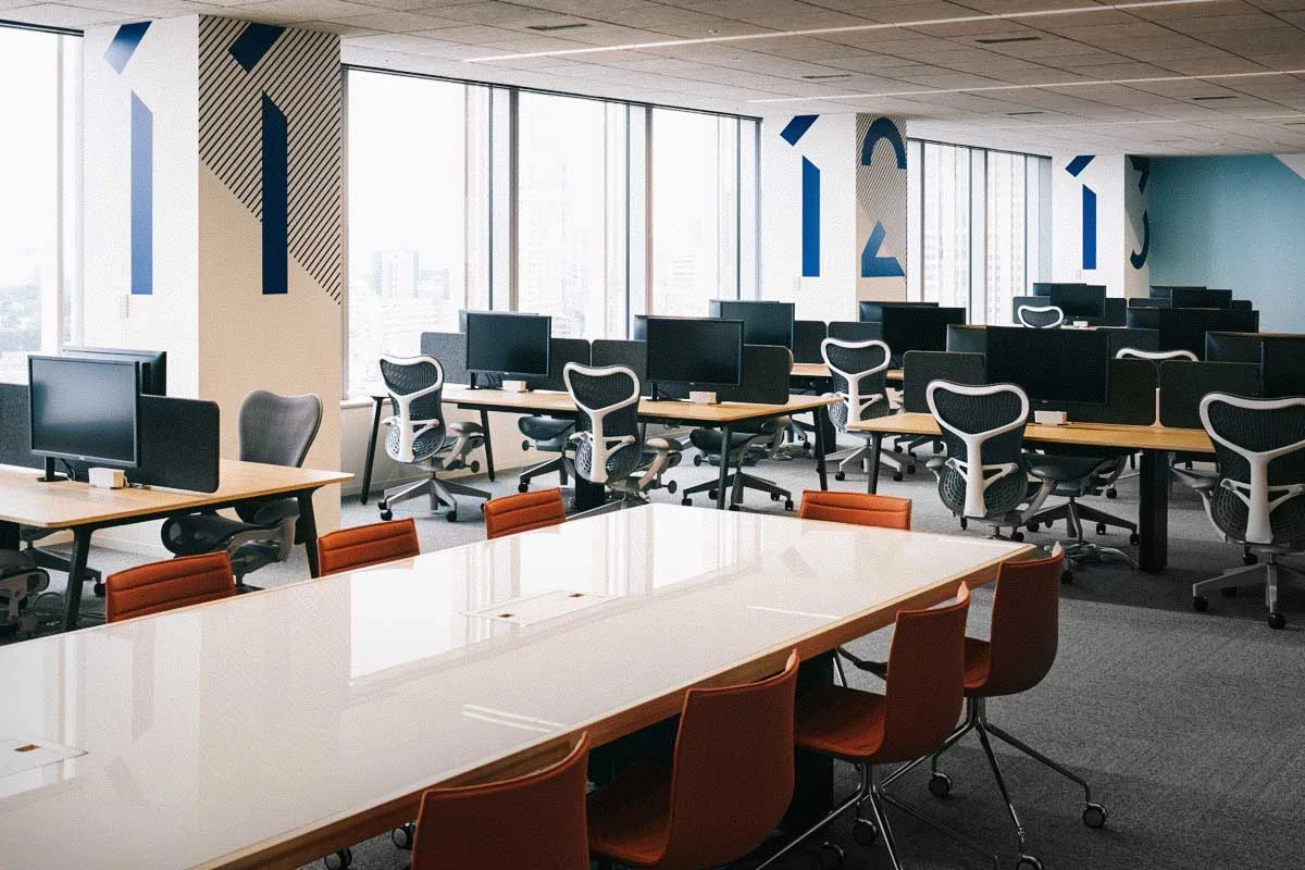 Empty modern office with arrange of computer desks and chairs by large windows, featuring a long conference table with orange chairs in the foreground.