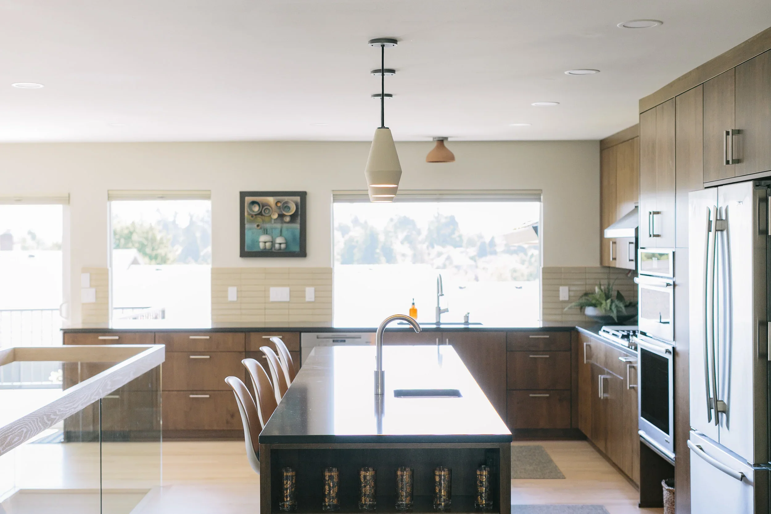 Modern kitchen with wooden cabinets, black countertops, a central island with bar stools, stainless steel appliances, and large windows letting in natural light.