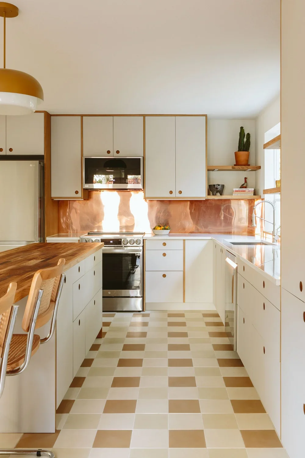 A modern kitchen with white cabinets, wood accents, a checkered beige and brown floor, and copper backsplash. Contains a stove, microwave, refrigerator, sink, and open shelves with plants and decor.