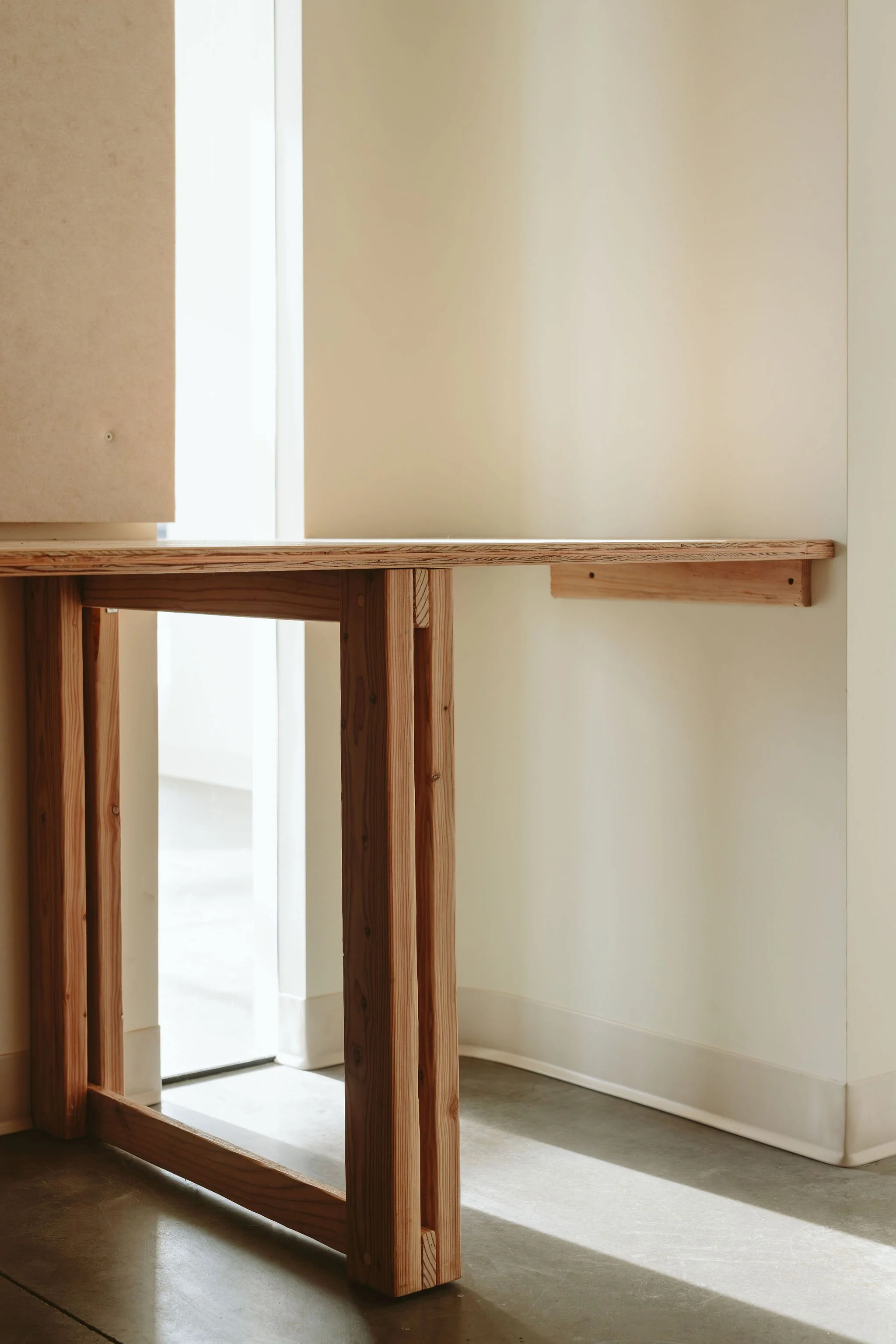 Interior of a room with a wooden table and a small wood shelf attached to the wall, with sunlight coming through a window.
