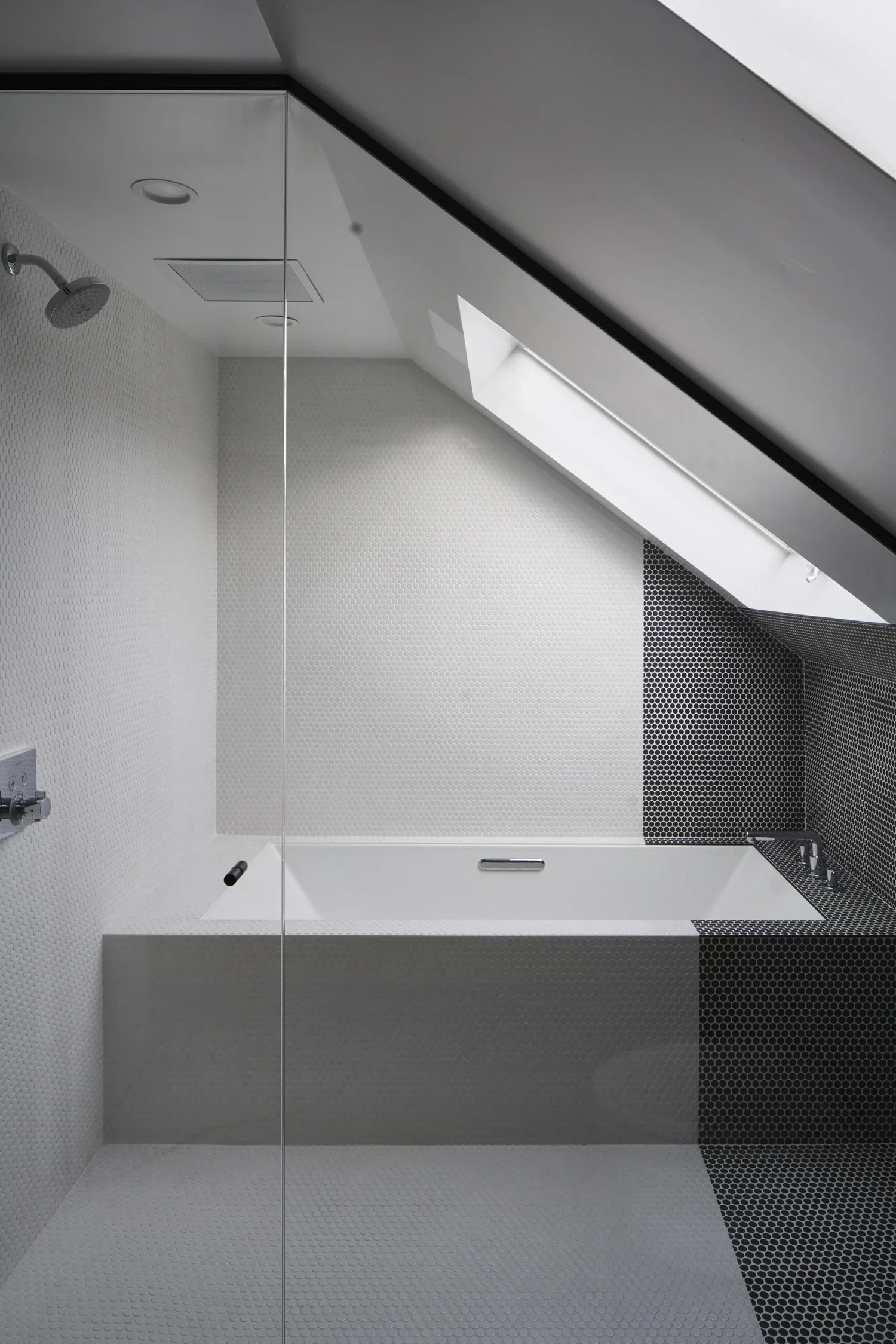 Modern bathroom shower area with a white bathtub, gray and black mosaic tiles, a skylight window, and a ceiling shower head.