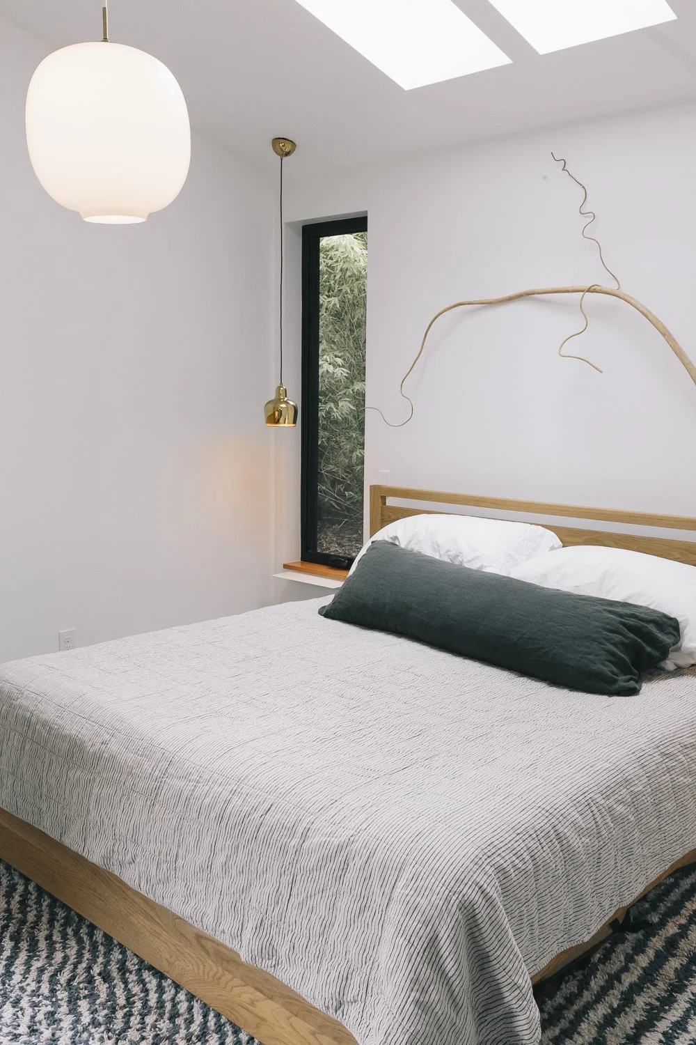 A minimalist bedroom with a bed, a large paper lantern ceiling light, a gold pendant light, a black framed window, and a decorative branch on the wall.