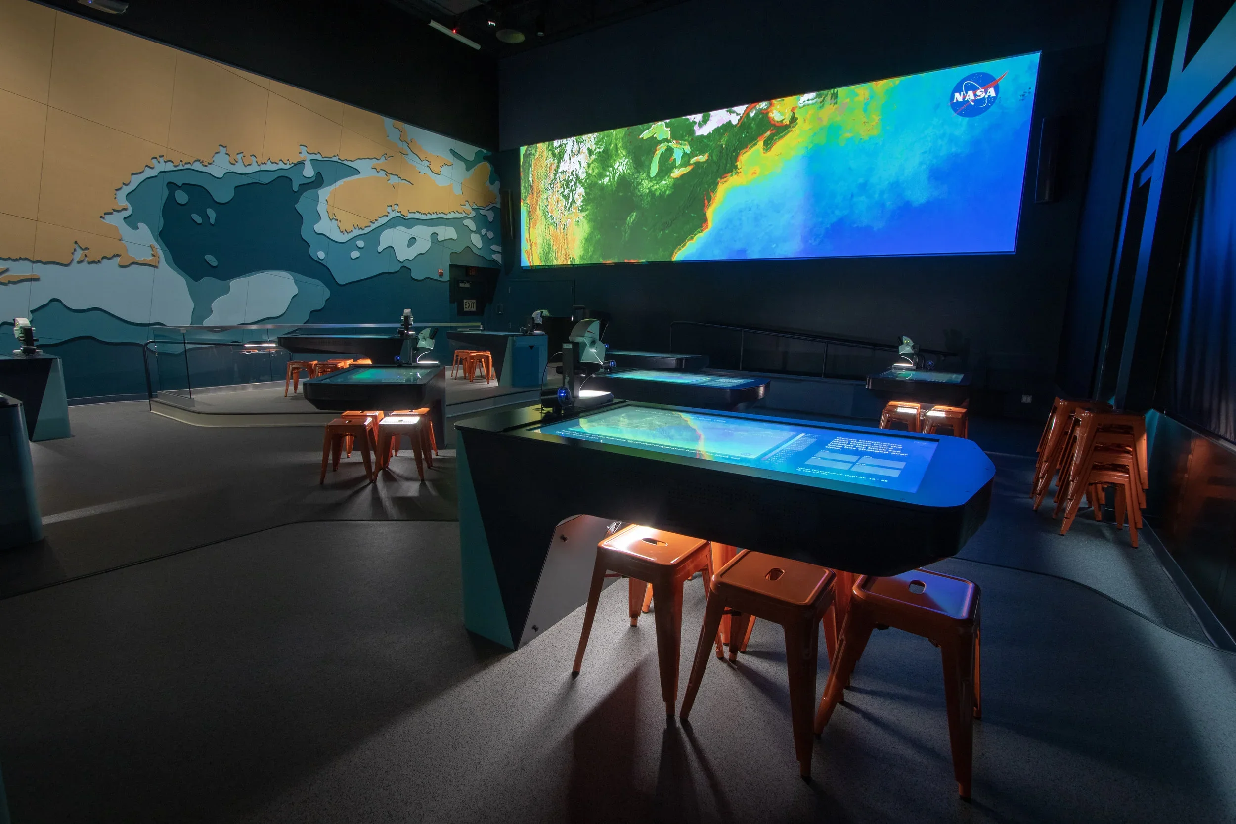 Interior of a NASA exhibit room with large digital screens displaying a map and satellite imagery, decorated with a world map on the wall and orange stools around interactive tables.