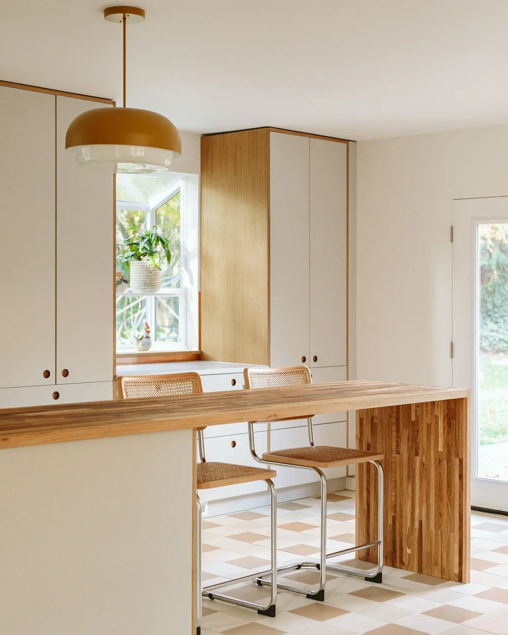Minimalist kitchen with white cabinets, a window with a plant, wooden accents, a wooden kitchen island, and two barstools with wicker seats. There is a large pendant light hanging from the ceiling.