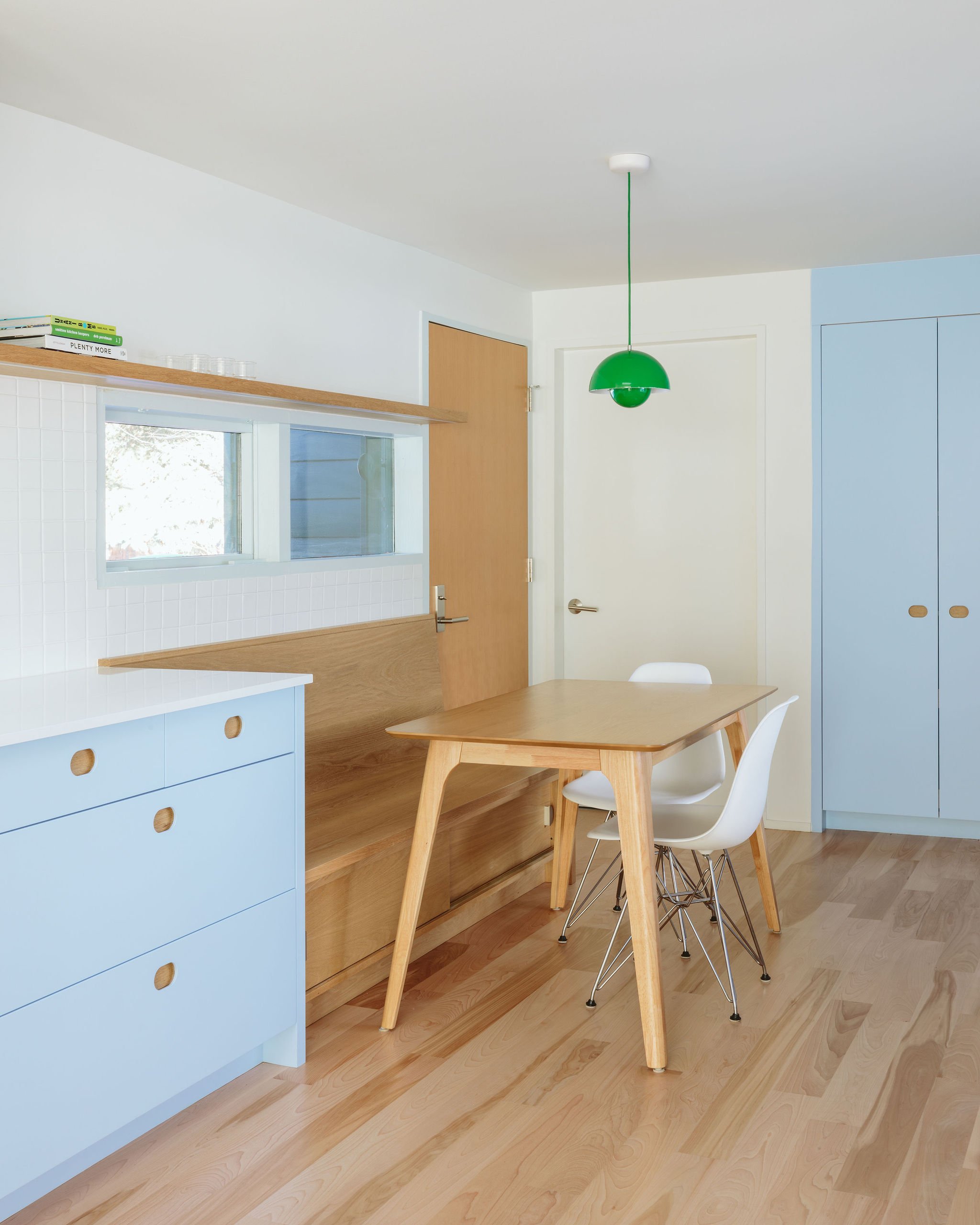 Modern dining area with a wooden table, white and blue chairs, light wood flooring, a blue cabinet, a wall with a window, a wooden door, a green pendant light, and built-in blue storage cabinets.