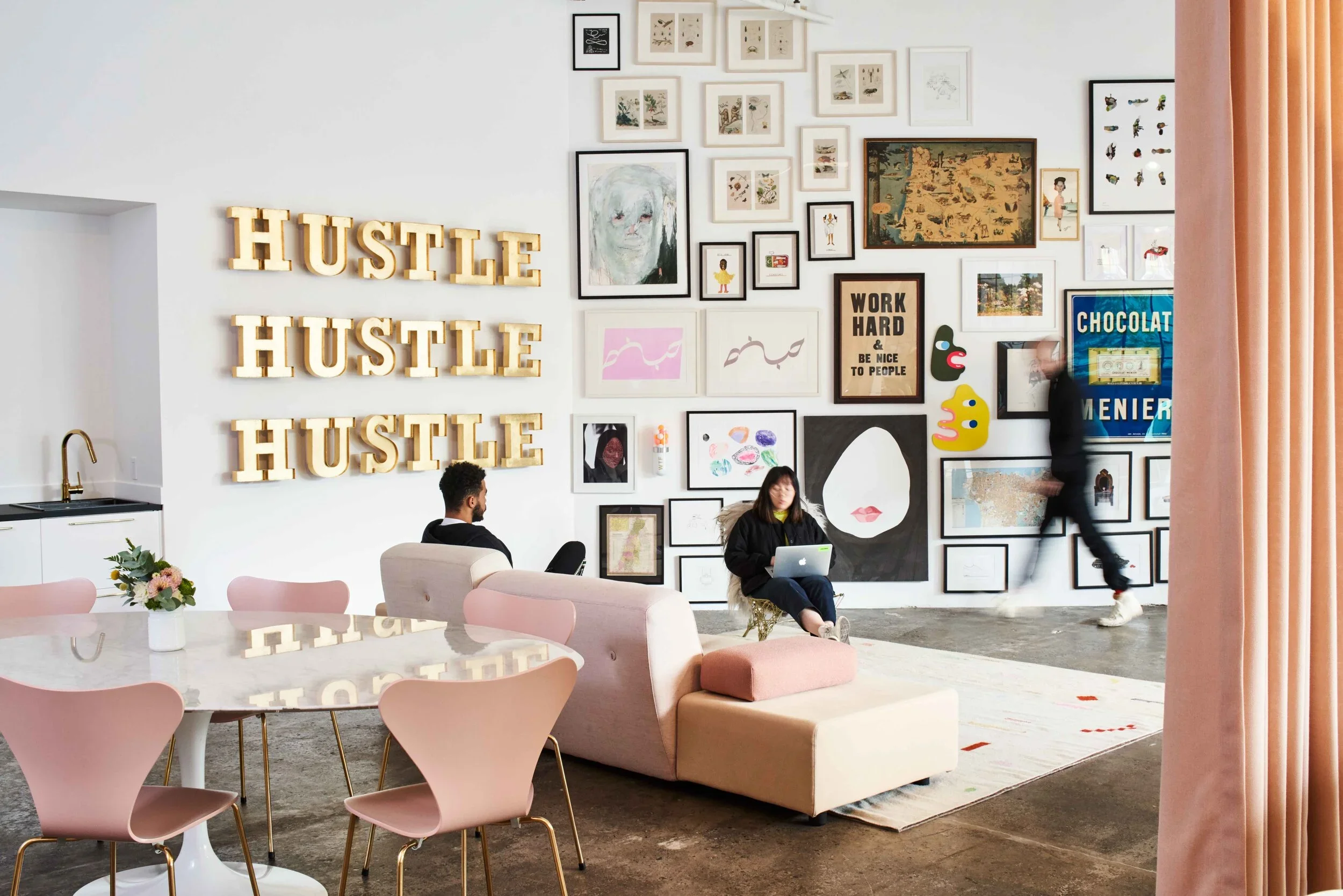 A modern, stylish room with a gallery wall featuring framed artwork and colorful decorations, large wooden letters spelling 'HUSTLE' three times, a pink and beige sofa with a woman working on a laptop, a man sitting nearby, a round table with pink chairs and a small flower arrangement, and a person walking past the wall.