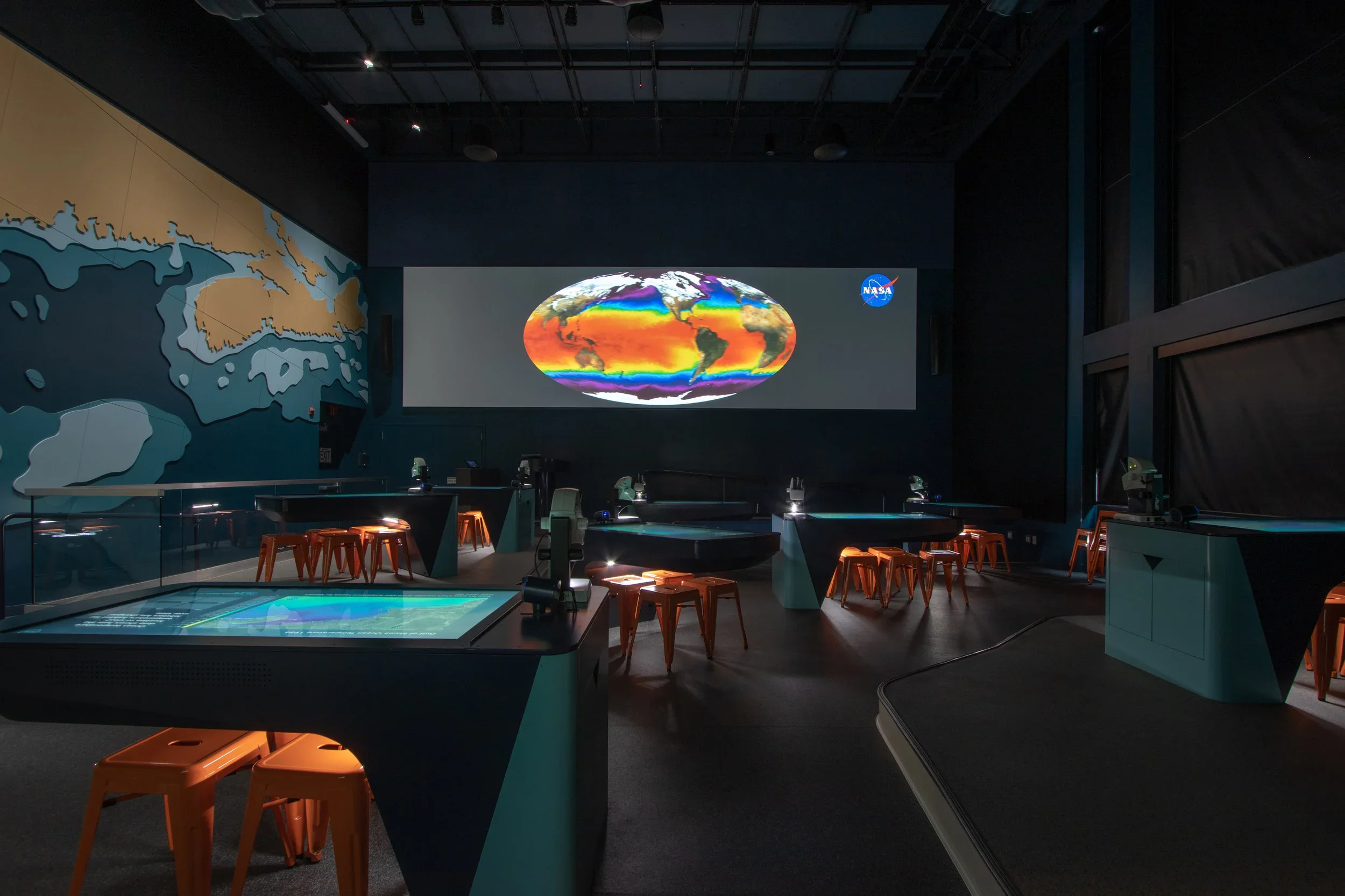 A science exhibit room with a large shopping-sized table with microscopes, orange stools, and a world map on the wall. A large screen displays a colorful map of Earth, with the NASA logo in the corner.