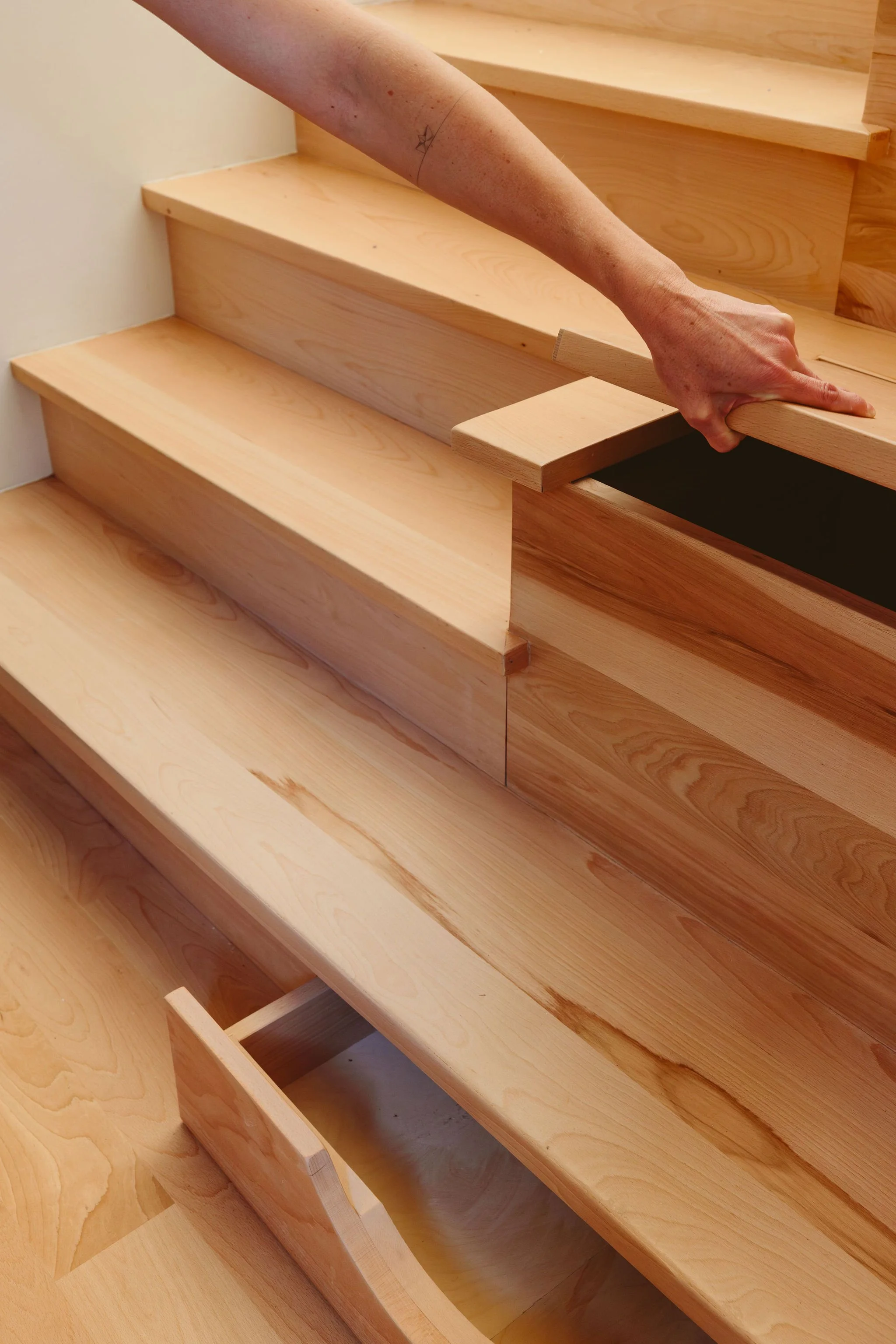 A person installing or inspecting wooden stairs or a staircase, with a focus on the hand holding a piece of wood. The staircase is made of light-colored wood with visible grain patterns.