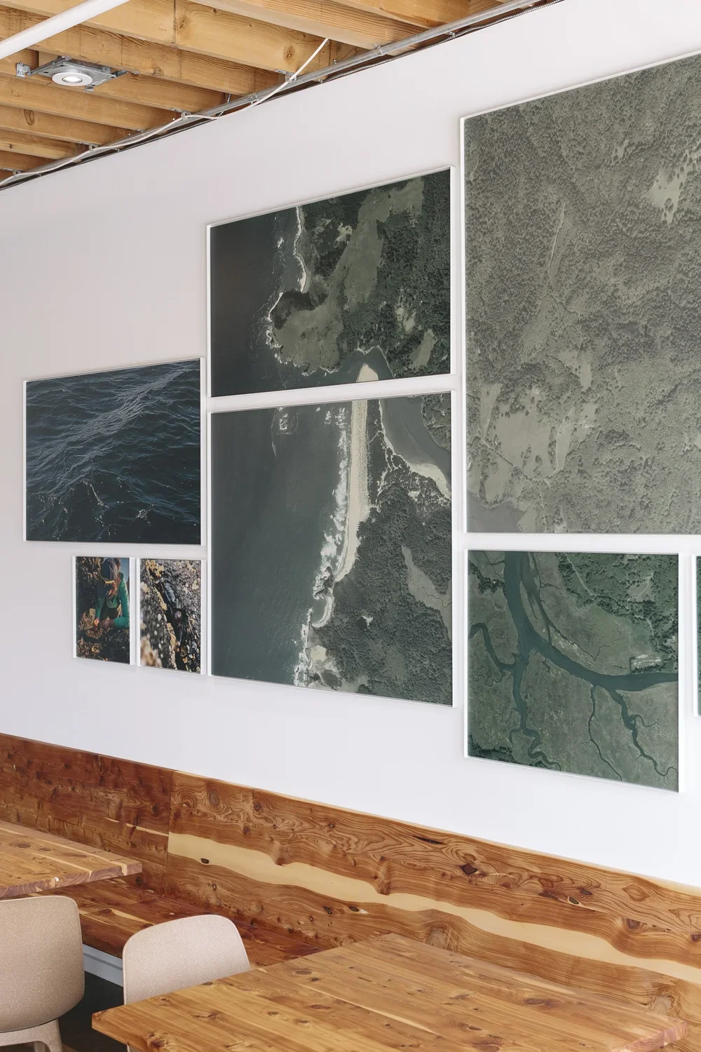 Gallery display of aerial landscape photographs on a white wall, featuring rivers and coastal areas, in a room with wooden tables and chairs.