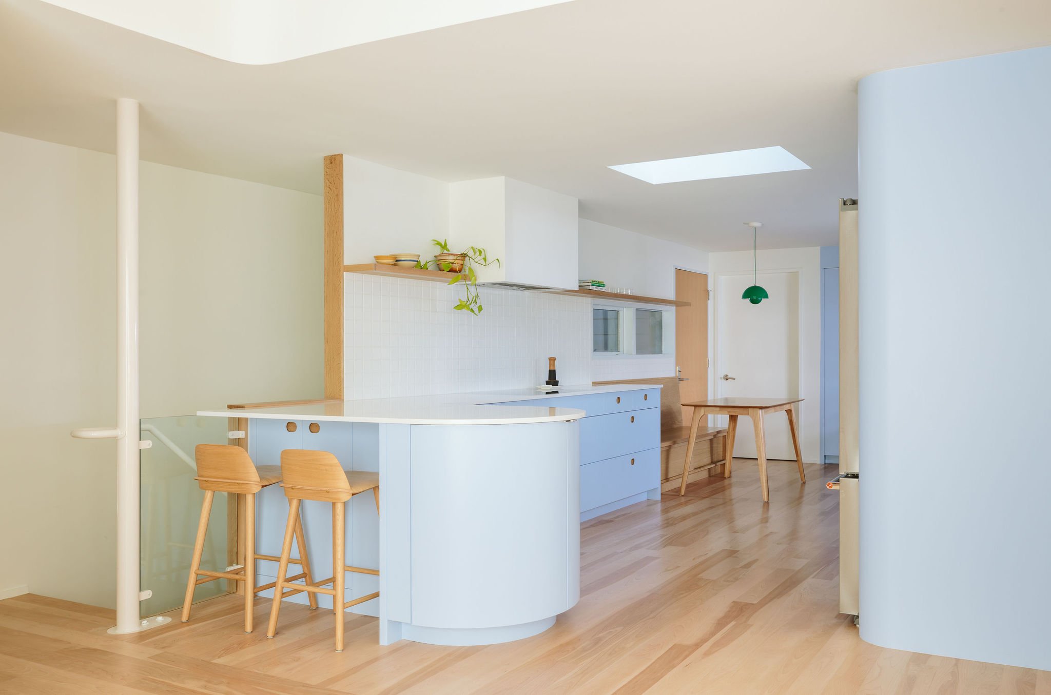 Modern kitchen with light blue cabinets, wooden accents, and light wooden flooring. There are two wooden bar stools at a white curved kitchen island, and a small table with matching wooden legs. The ceiling has skylights and a green pendant light. The background features a doorway and window, with a minimalistic design.