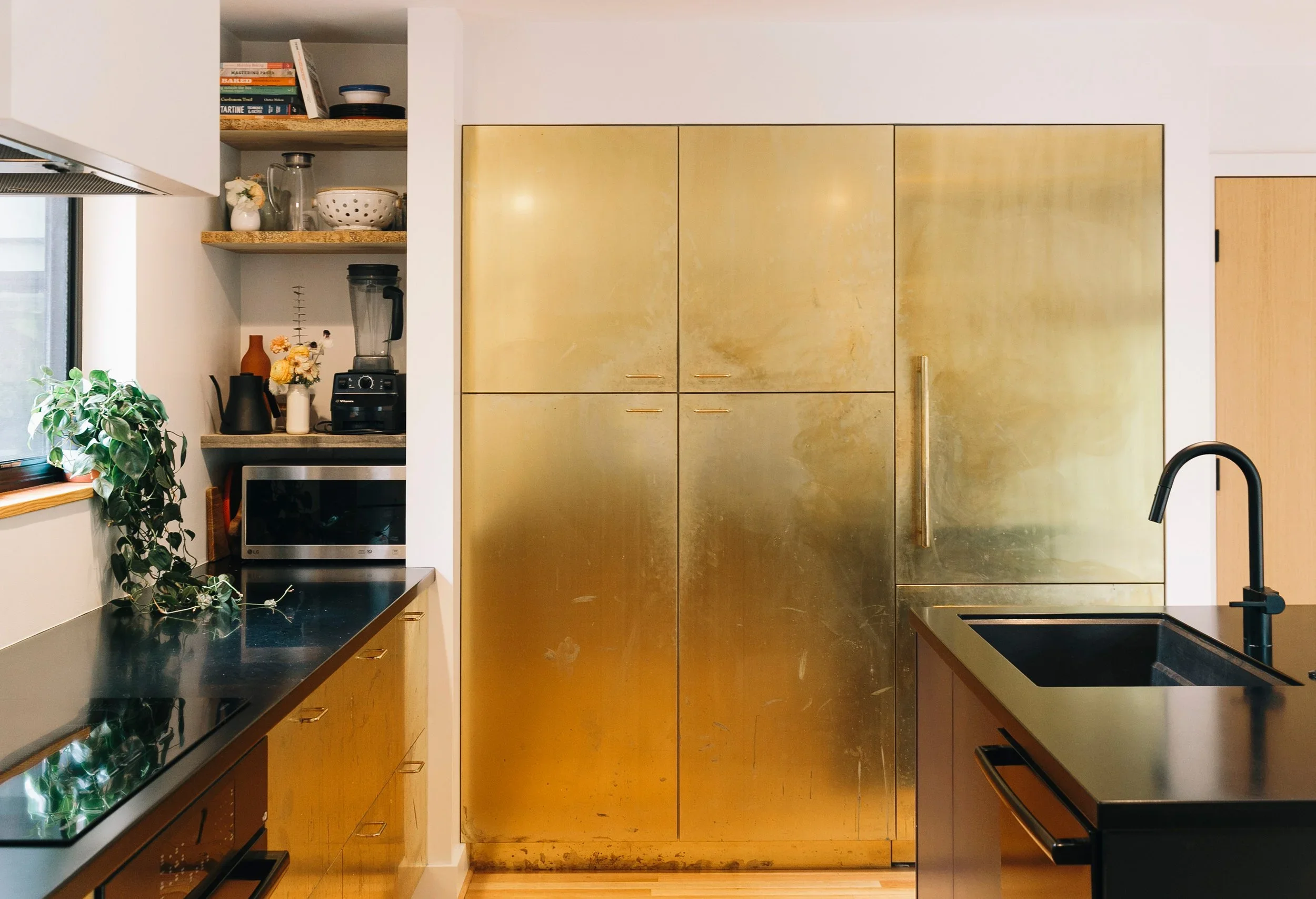 Kitchen with black countertops, wooden cabinets, a large gold-colored fridge, black sink with faucet, microwave, open shelves with books and bowls, blender, plants, and a window