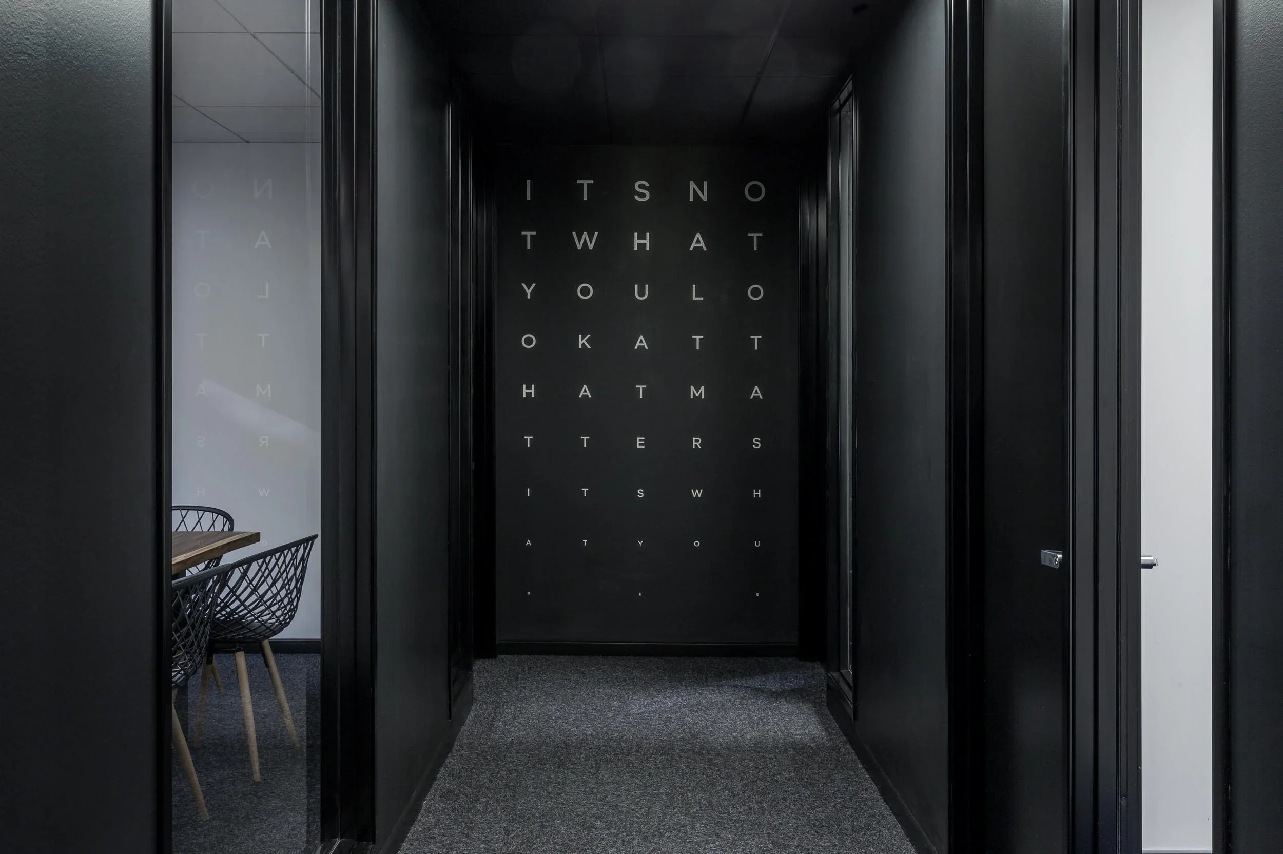 Black hallway with walls inscribed with alphabet letters and a conference room on the left with chairs and a table.