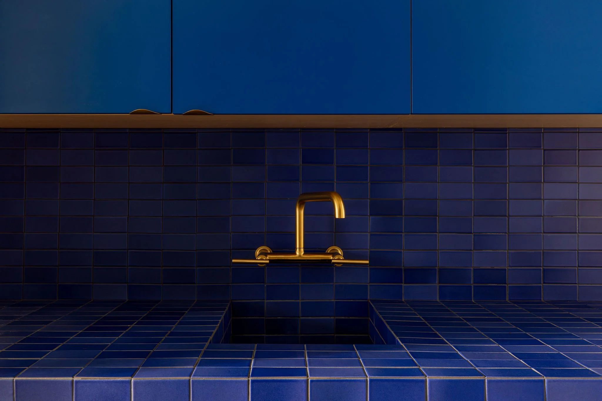 Blue tiled kitchen wall with a gold faucet and sink. Upper cabinets are also blue, with a wooden trim.