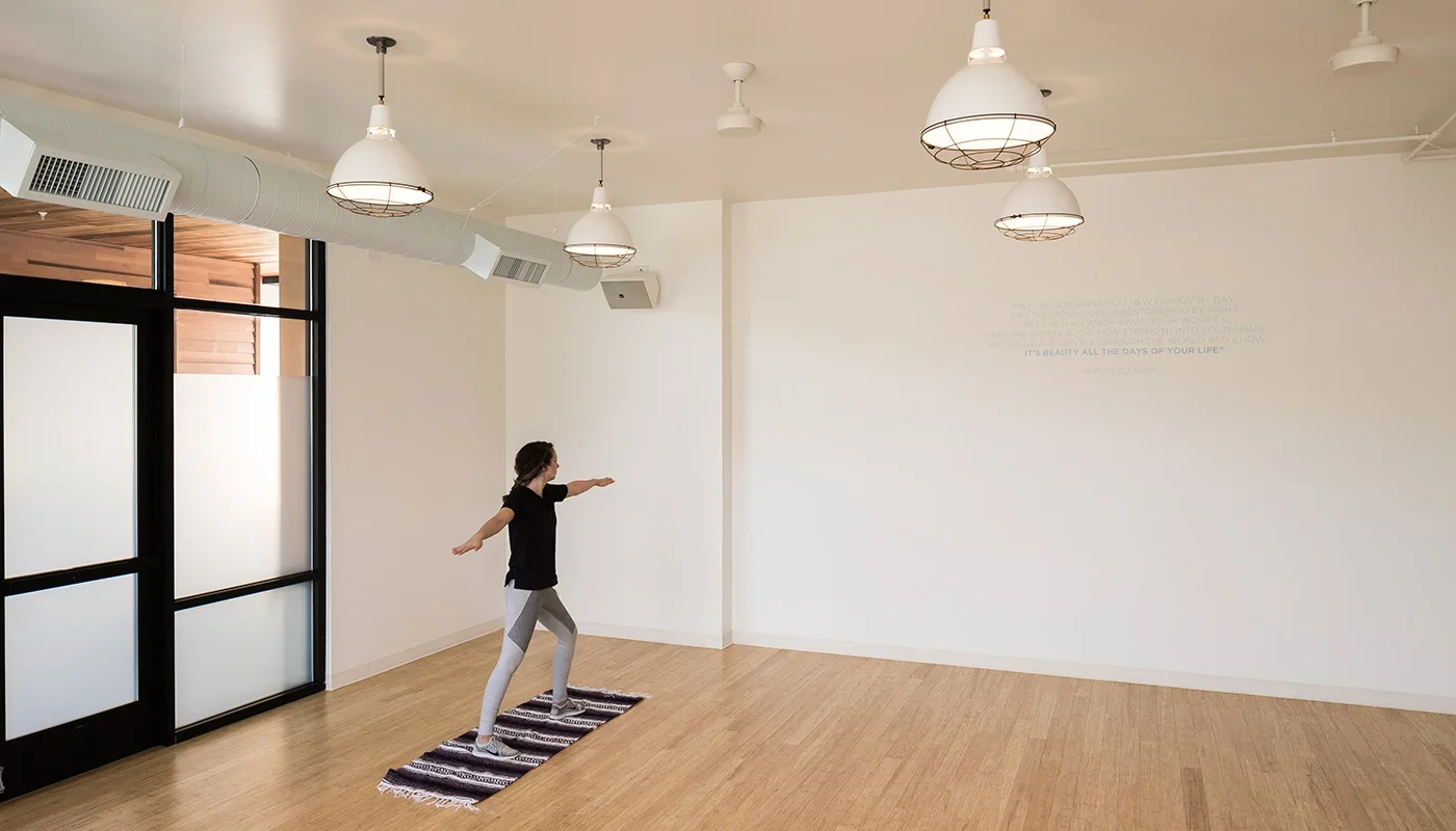 A young woman in workout clothes doing yoga or stretching on a small rug in a bright, empty room with white walls, wooden floor, and ceiling lights.