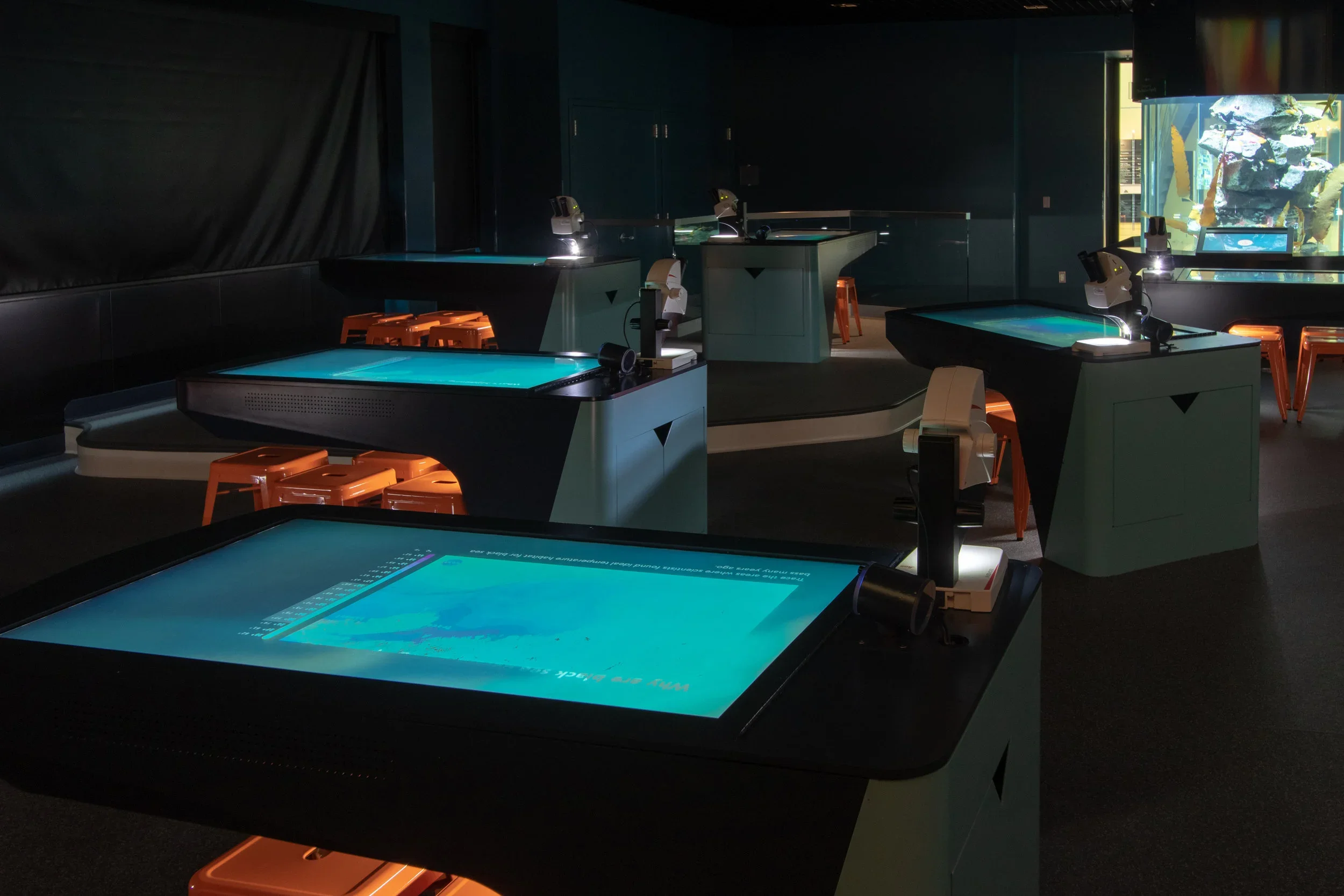 Empty planetarium or science exhibit room with several interactive touchscreen tables and microscopes, dimly lit, with a large aquarium or display visible through a window on the right.