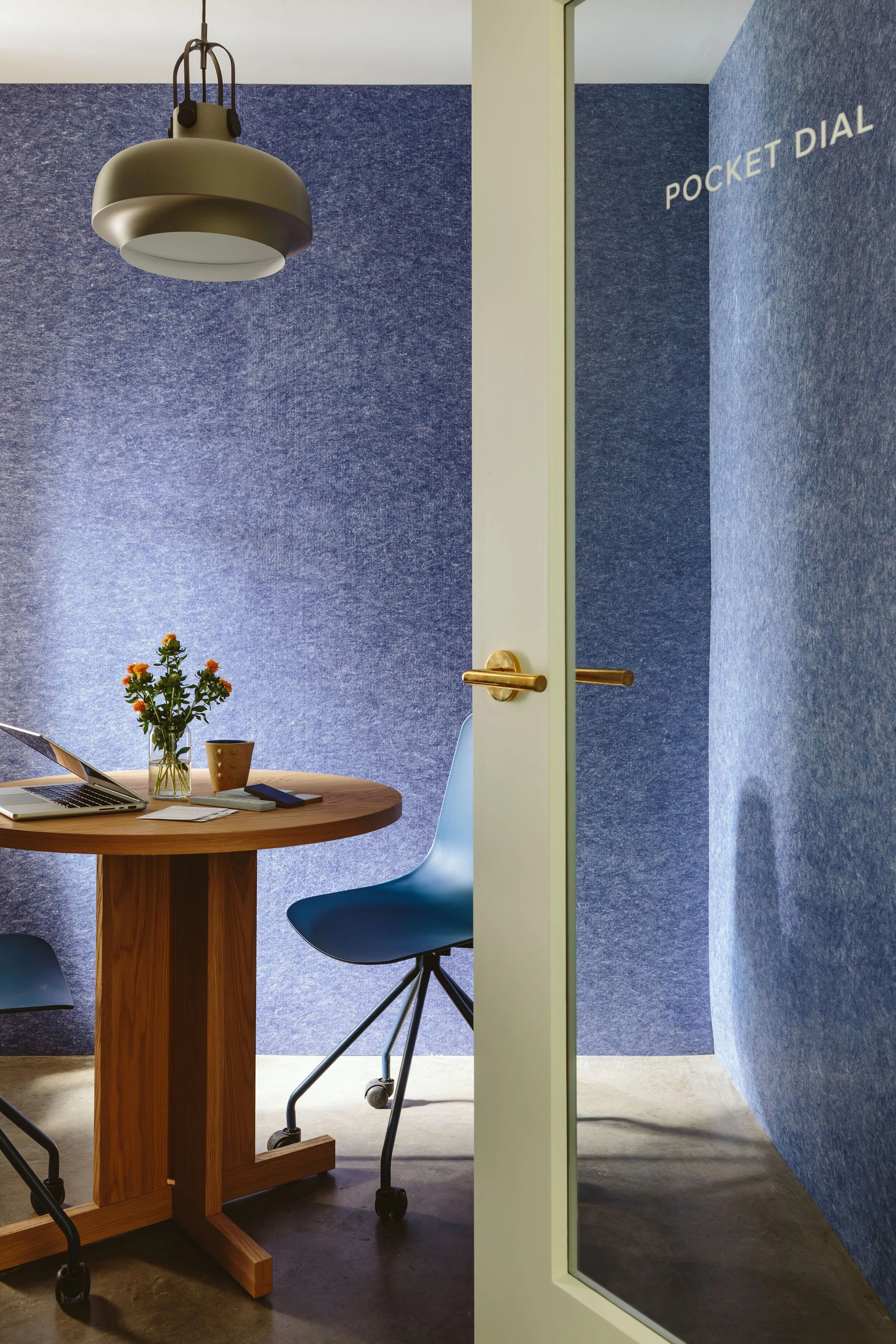 Partially open door leading to a small conference room with a round wooden table, a blue chair, a laptop, a laptop stand, a cup, a glass vase with flowers, and a hanging ceiling light. Blue upholstered walls and a sign that says 'POCKET DIAL' are visible.