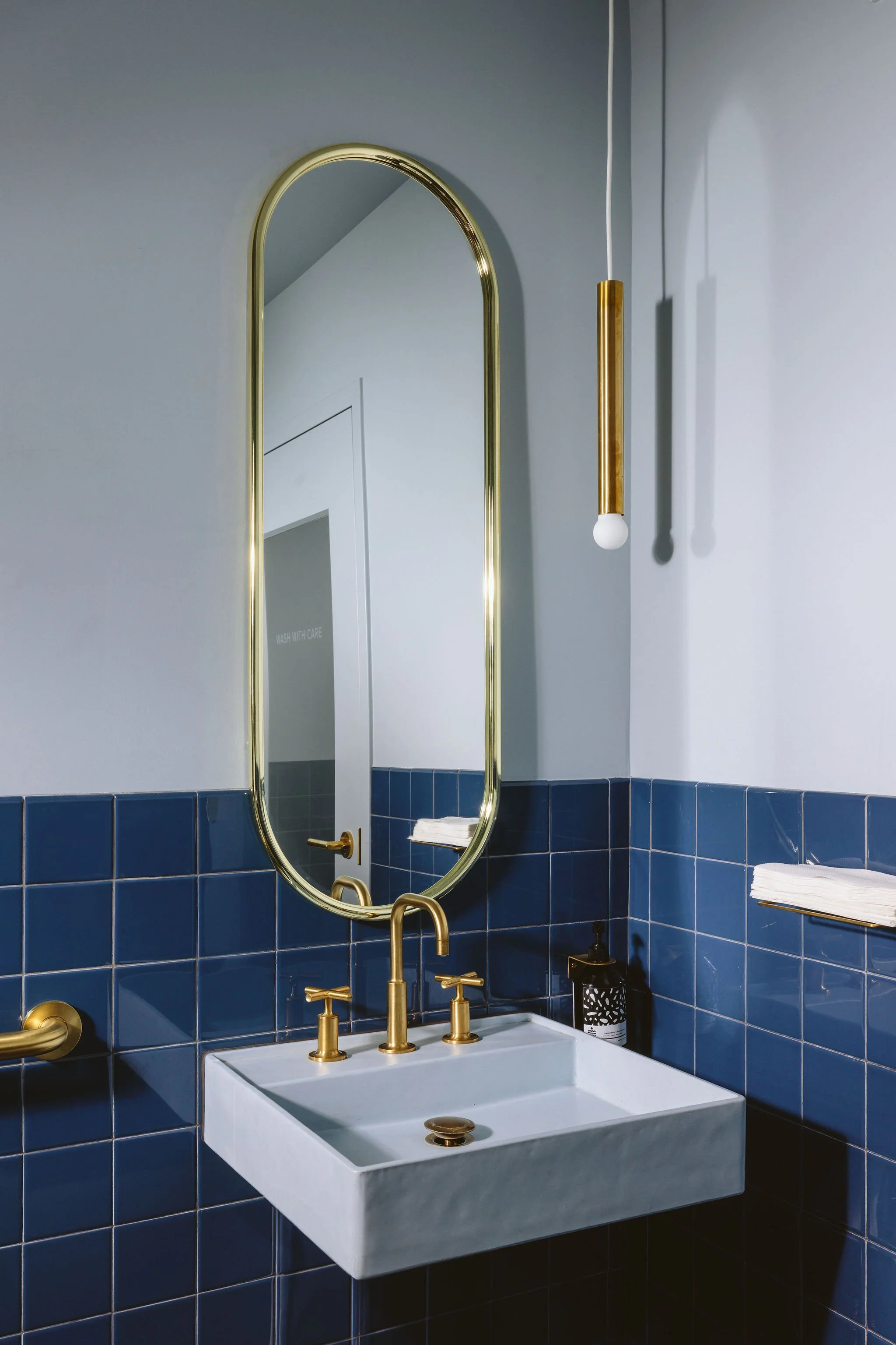 Modern bathroom with a white square sink, gold faucet, and blue tiled wall. An oval mirror with gold trim hangs above the sink, and a hanging gold and white light fixture is to the right. Towels are folded on a shelf to the right.