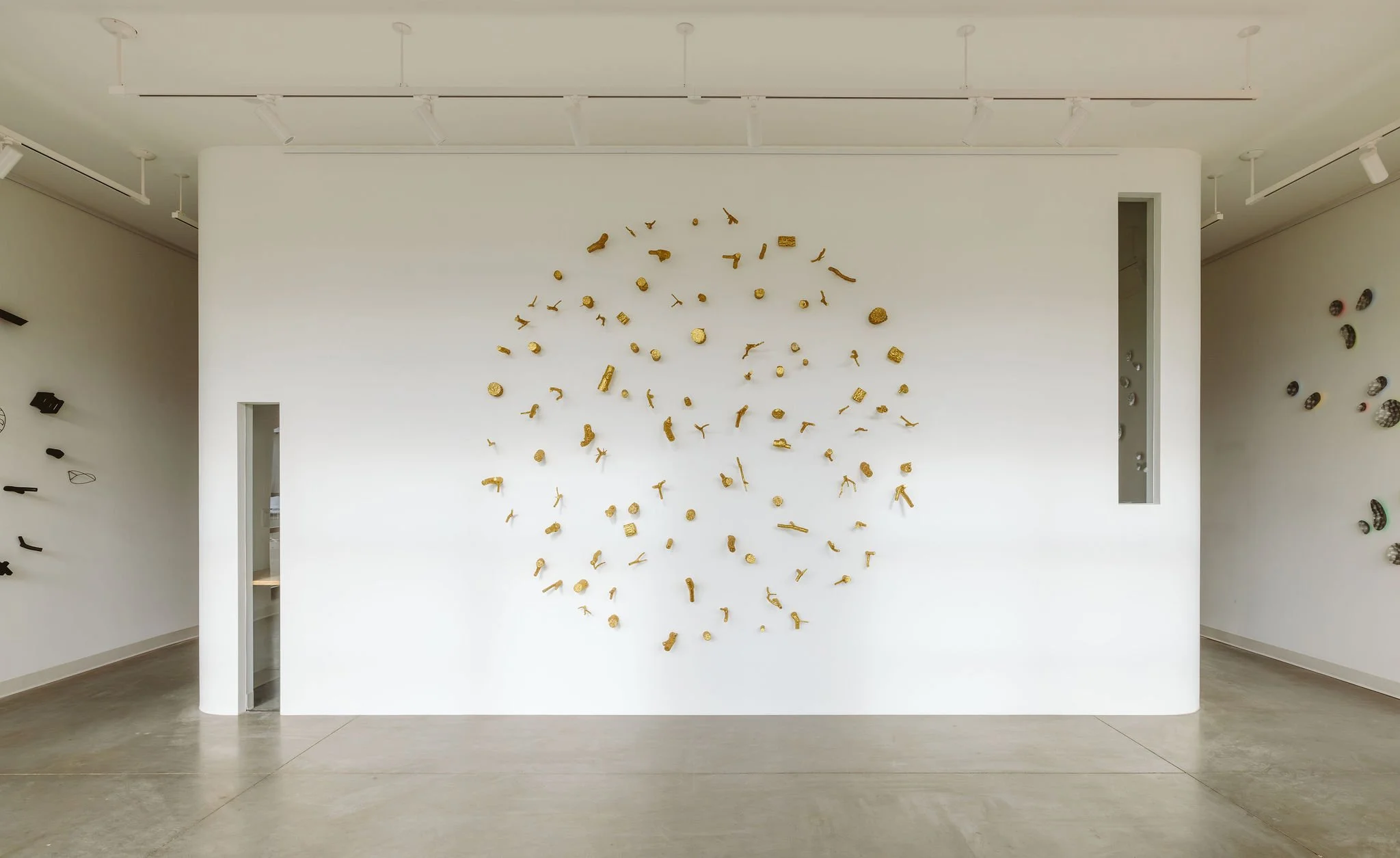 Art installation on white gallery wall featuring randomly arranged small, irregularly shaped, gold-colored objects.
